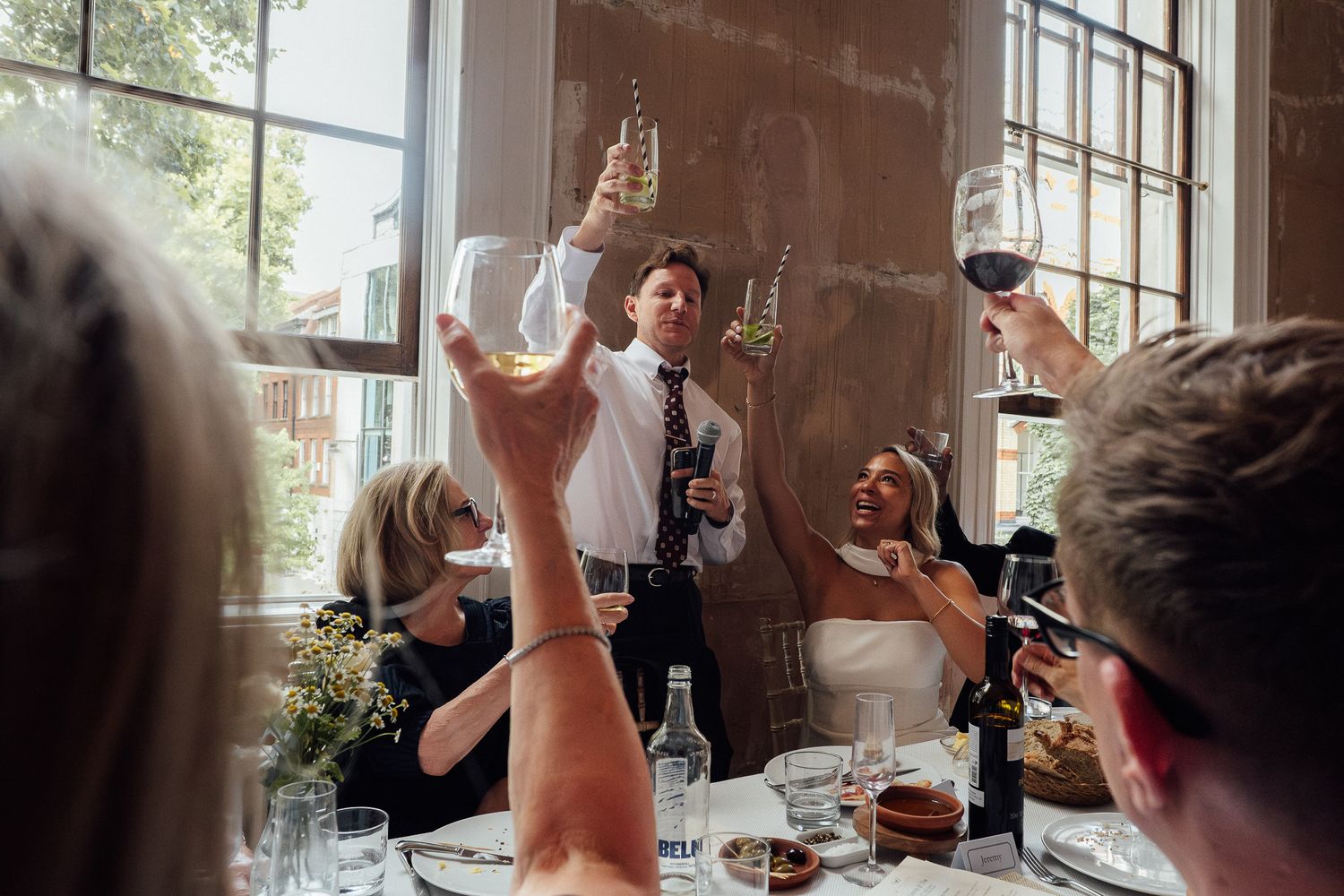 Guests raise wine glasses in a festive toast around a dining table as a man in a vest and tie speaks in a bright, sunlit room.