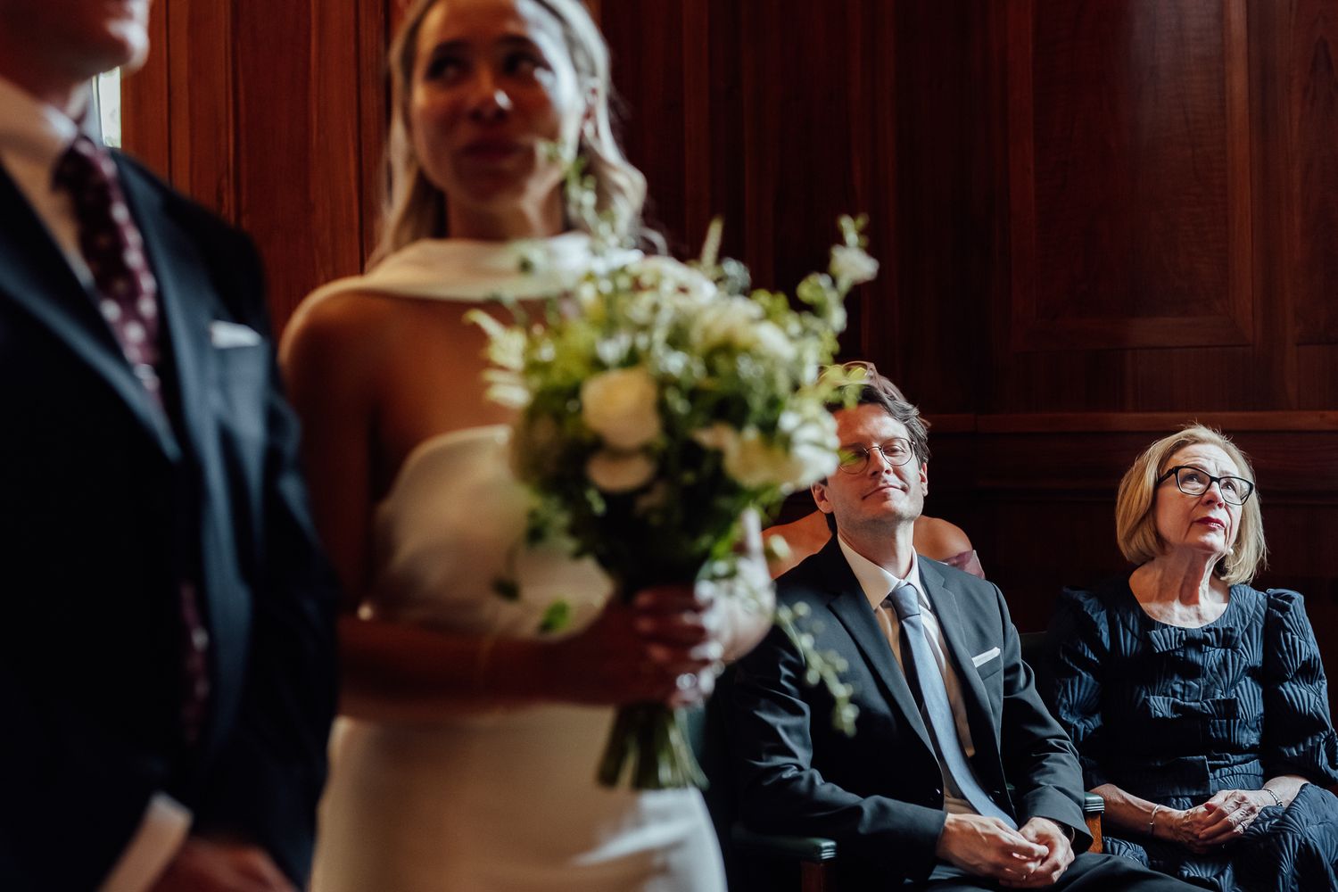 Bride in a white dress holds a white floral bouquet during a wedding ceremony while seated guests look on in a wood-paneled room.