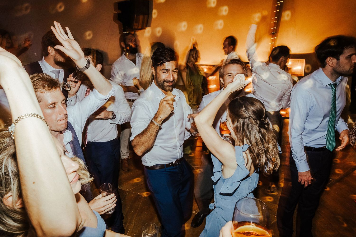 Energetic wedding guests dance and celebrate on a crowded dance floor with warm ambient lighting.
