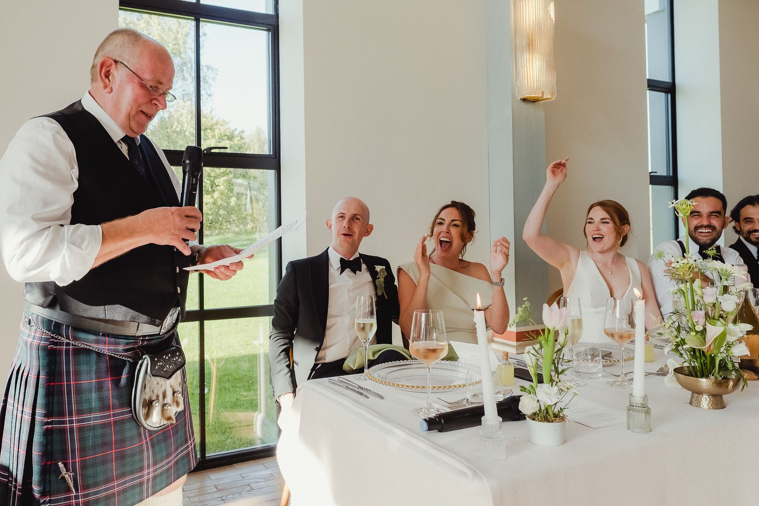Wedding reception guests laugh and react during emotional speech at elegant venue with large windows.