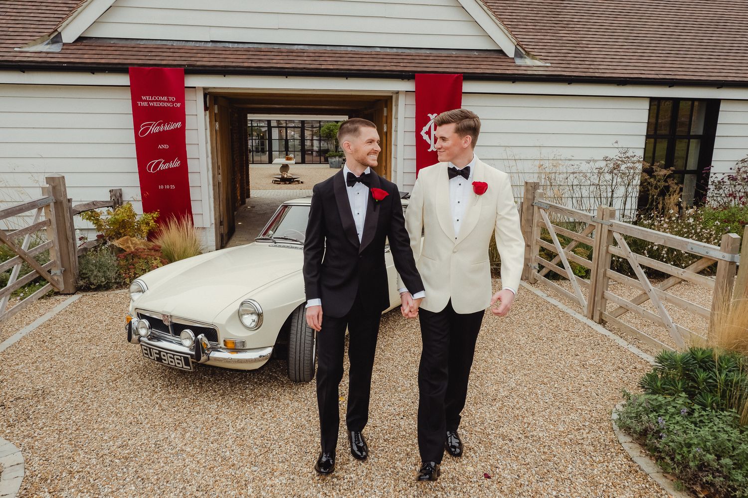A wedding party walks together on a gravel path outside a building with classic cars visible in the background.