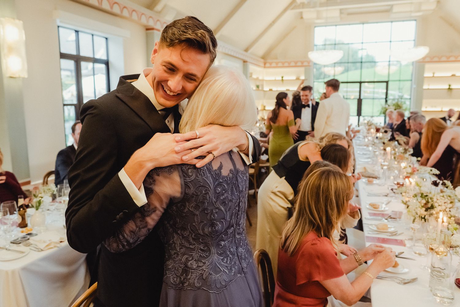A heartfelt embrace between two people during a wedding reception with dining tables and guests in the background.