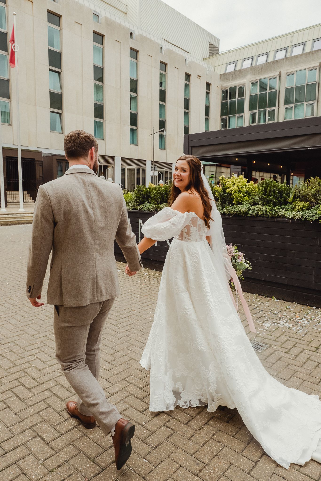 A couple walking on brick pavement outside a modern building, with the bride in a flowing white gown and long train.