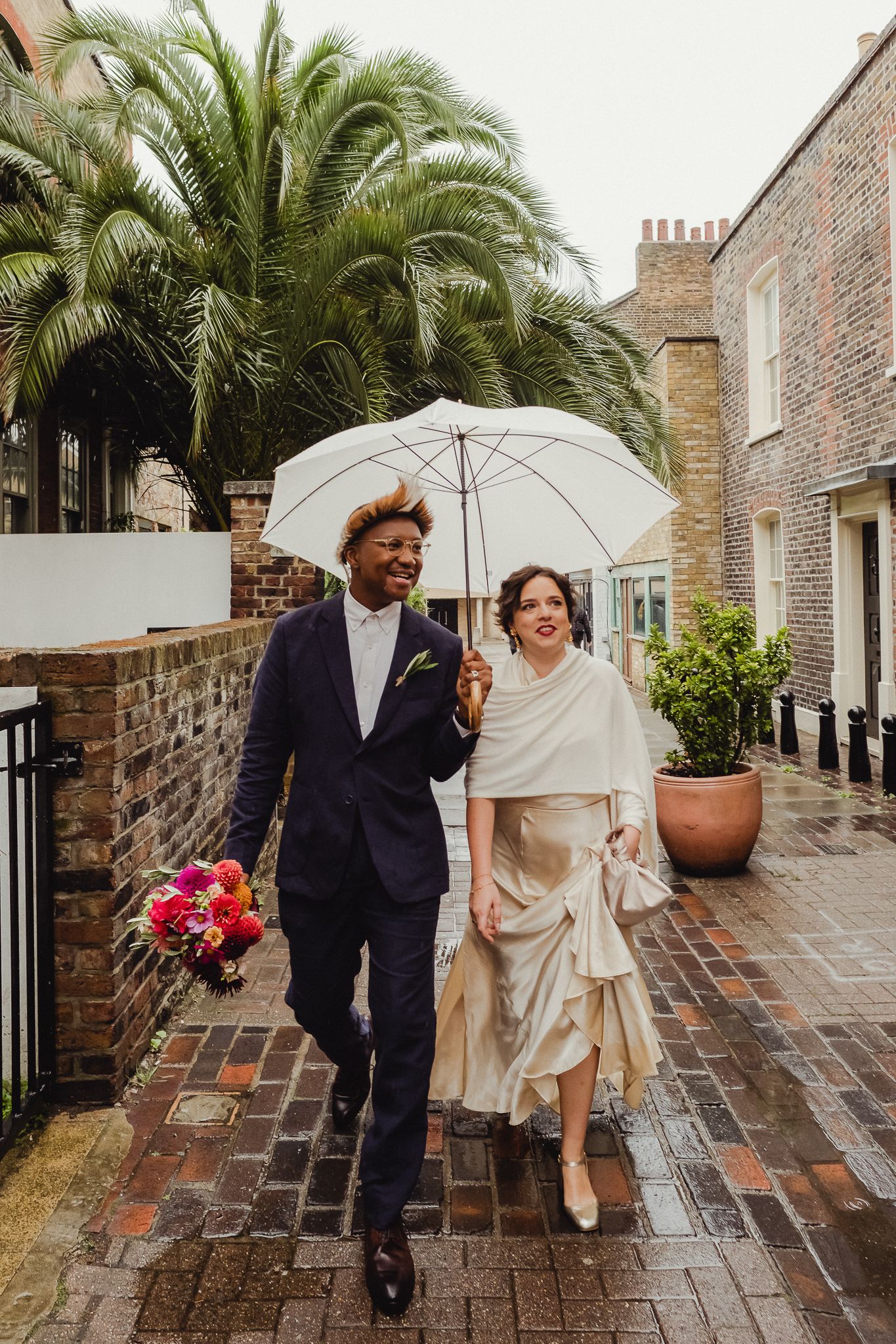 A couple walks together under white umbrellas on a rainy brick courtyard with palm trees in the background.