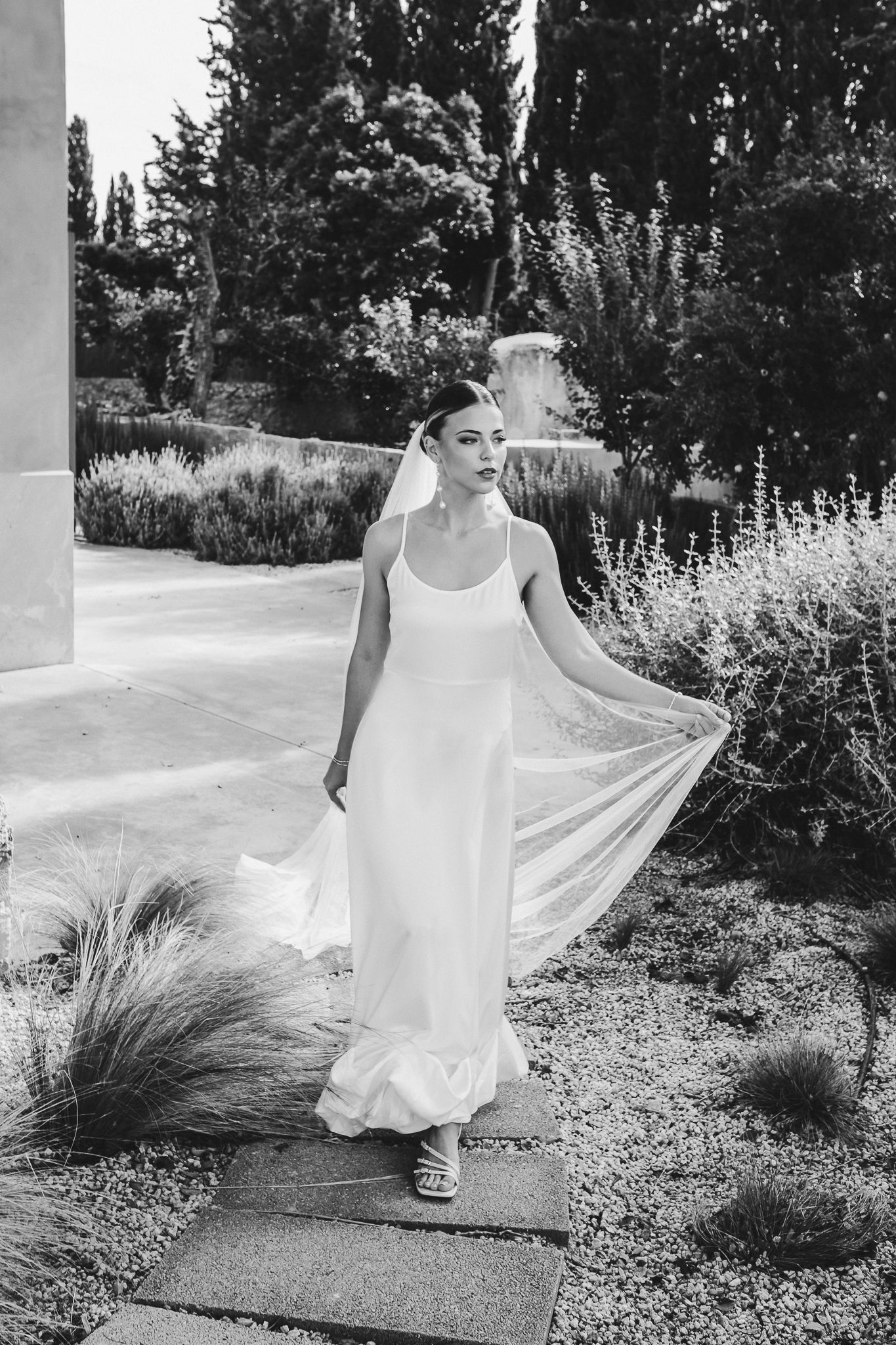 A graceful figure in a flowing white dress twirls through a garden setting with tall ornamental grasses and trees.
