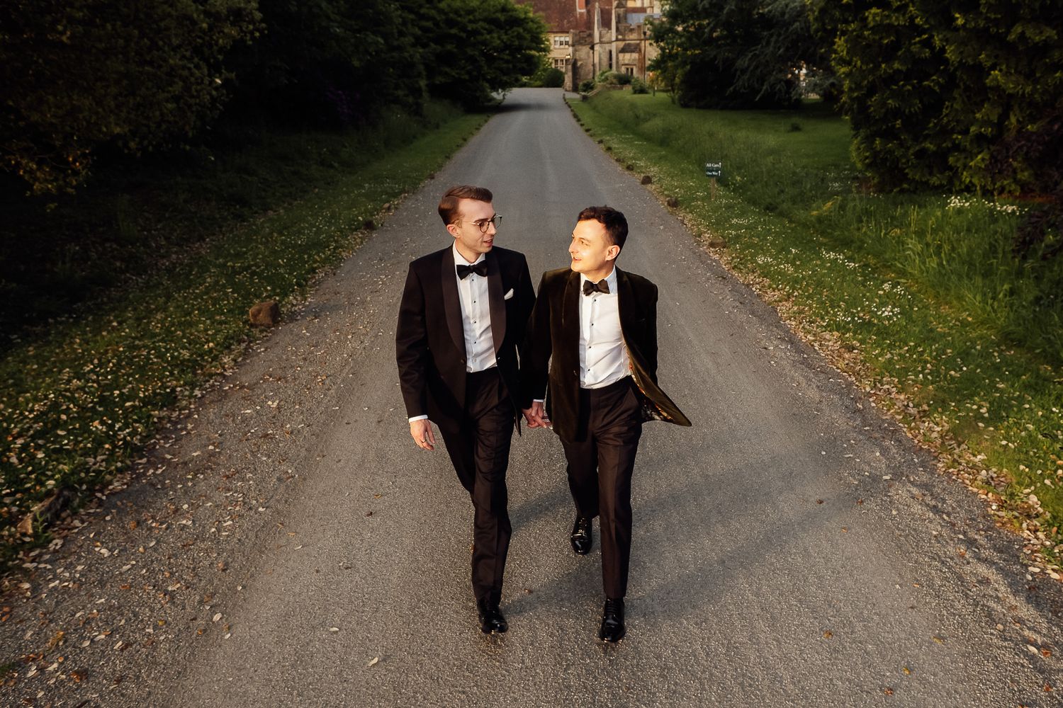 Two grooms in black tuxedos and bow ties walk hand in hand down a tree-lined path during golden hour.