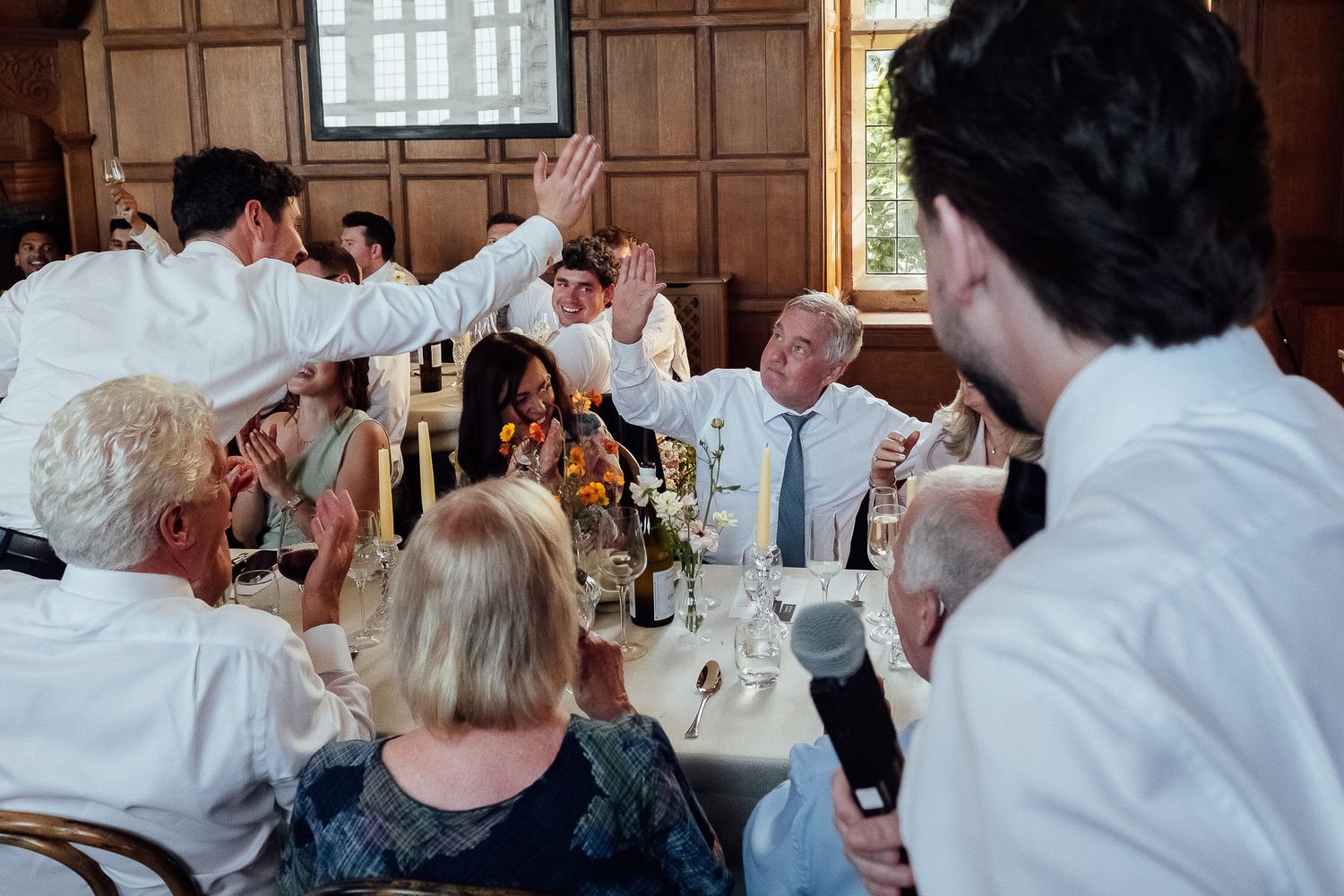 Wedding guests celebrate with high fives at a reception dinner in an elegant wood-paneled venue.