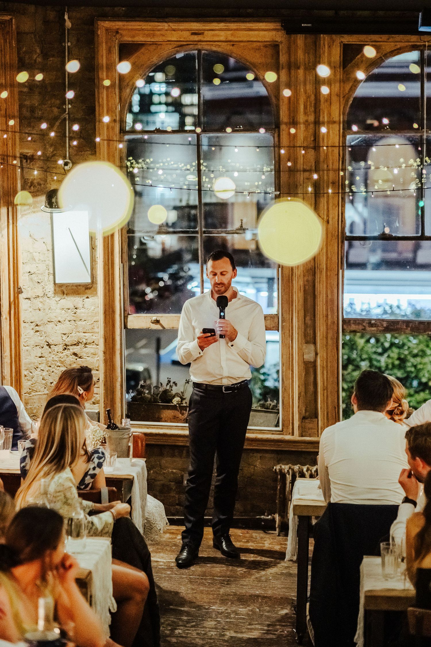 Wedding guests and servers mingle in an elegant rustic venue with glowing pendant lights and exposed brick walls.