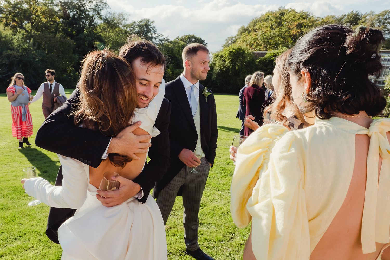 Wedding party members walk and laugh together on a grassy lawn during an outdoor celebration.