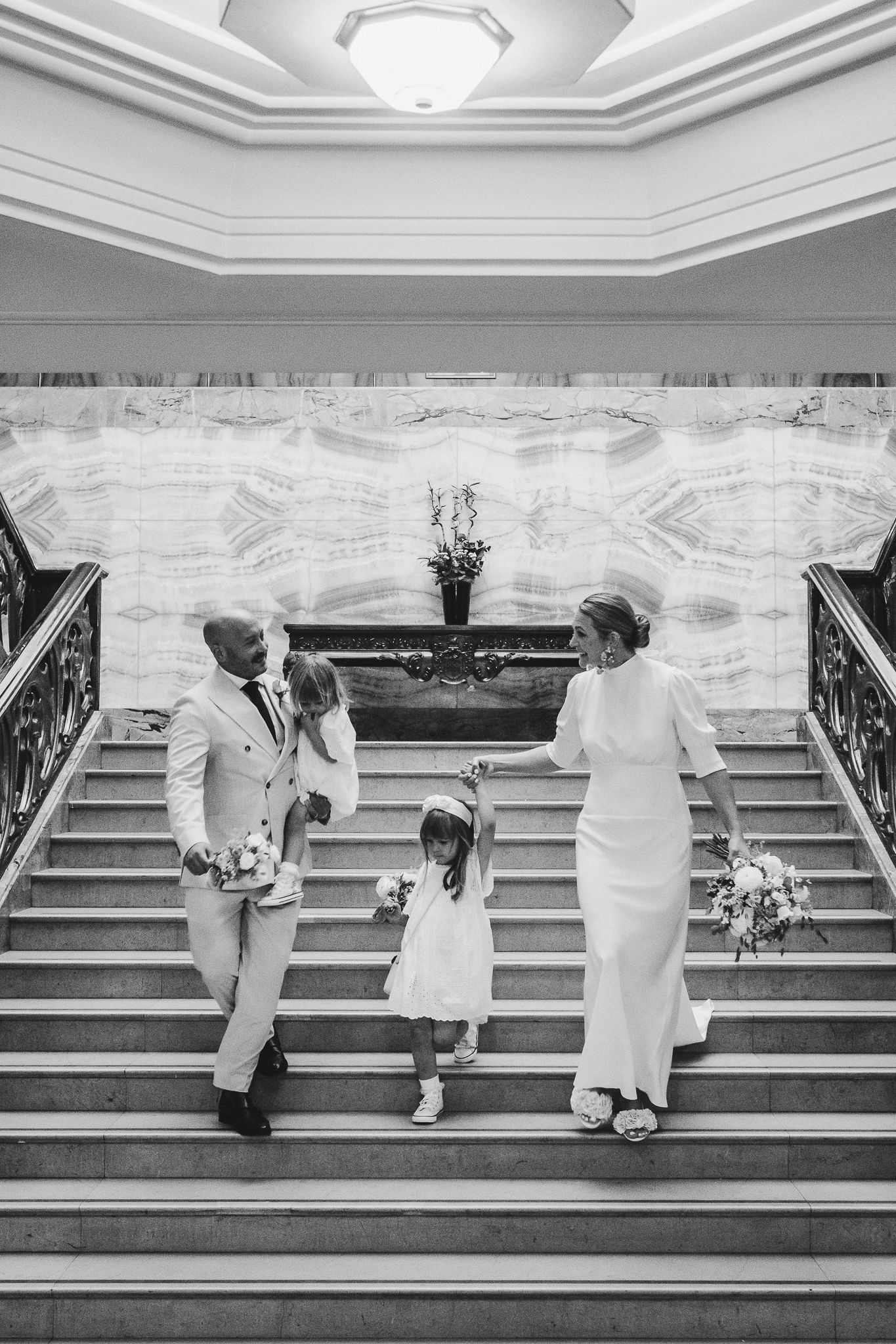 Black and white photo of a family in formal attire standing on an elegant staircase with marble walls and ornate railings.