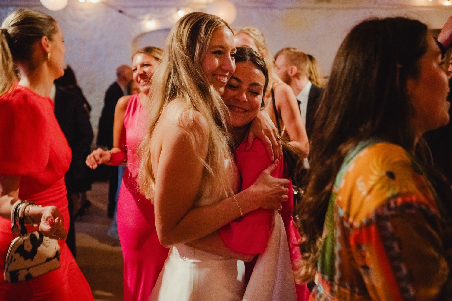 A group of party guests dancing and socializing on a dimly lit dance floor with vibrant pink and yellow dresses.