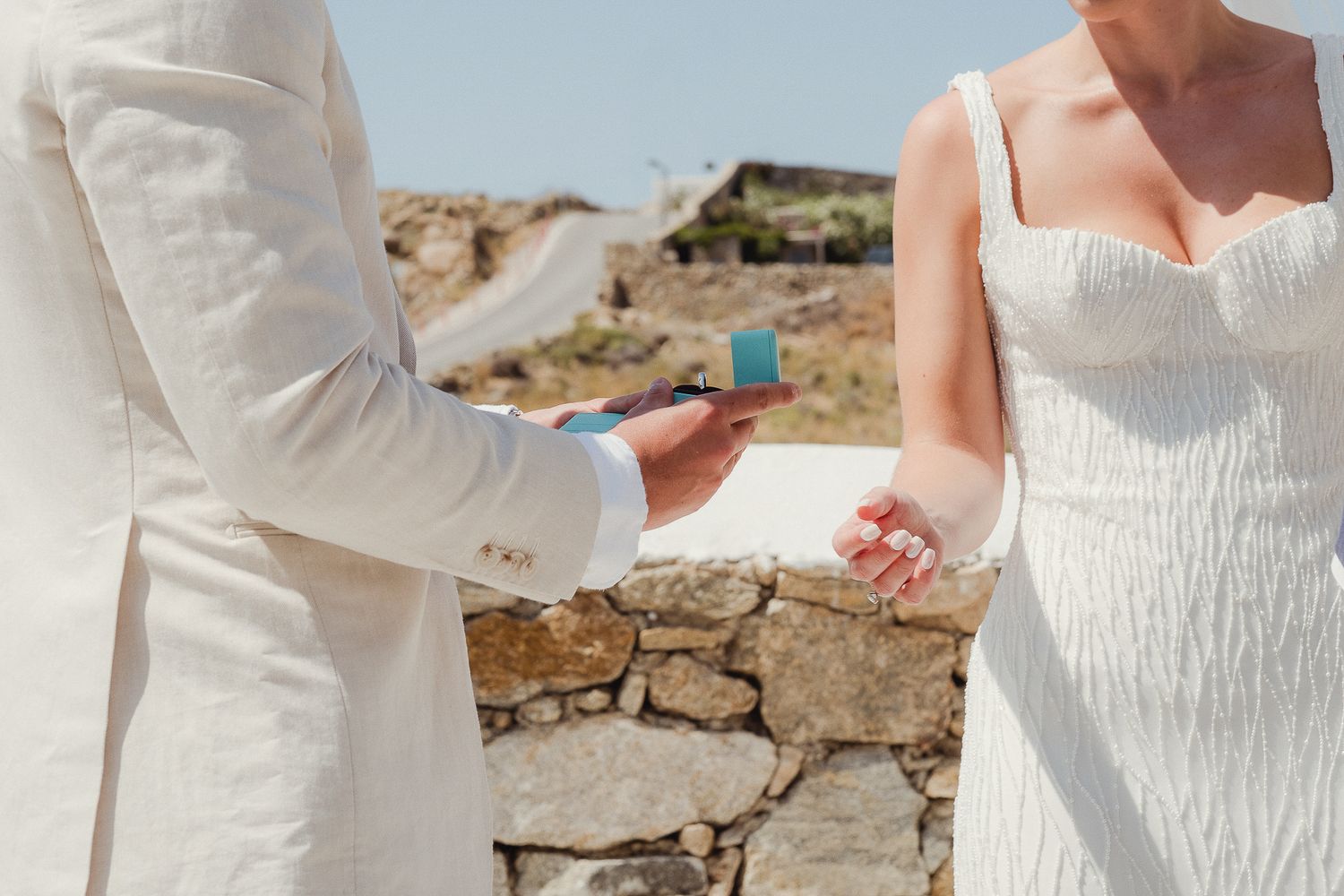 A group of people in white gowns and dresses gather on a rocky shoreline for a wedding celebration.