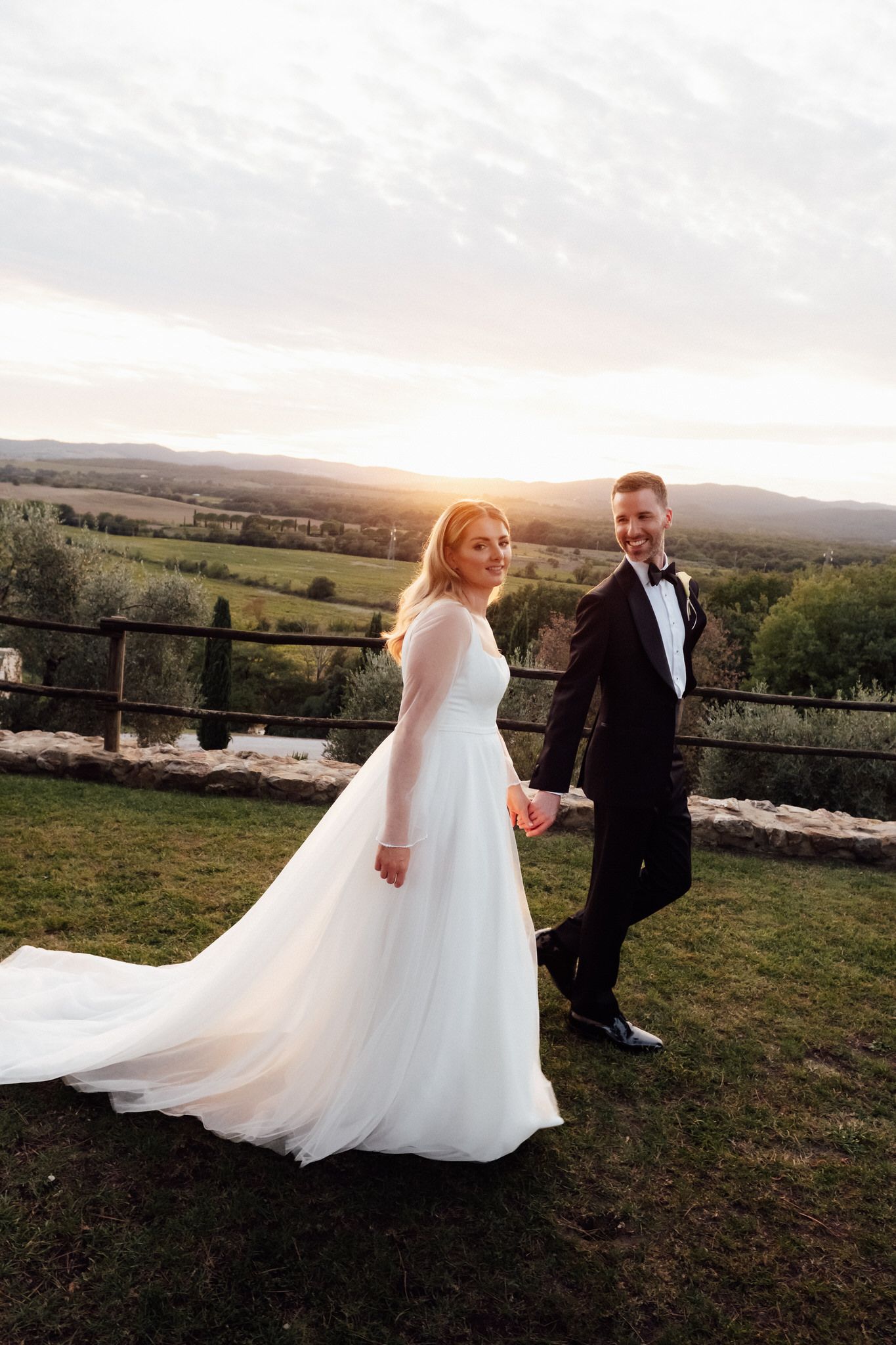 Bride in a flowing white gown and groom in a black tuxedo hold hands on a grassy hillside at sunset overlooking rolling countryside.