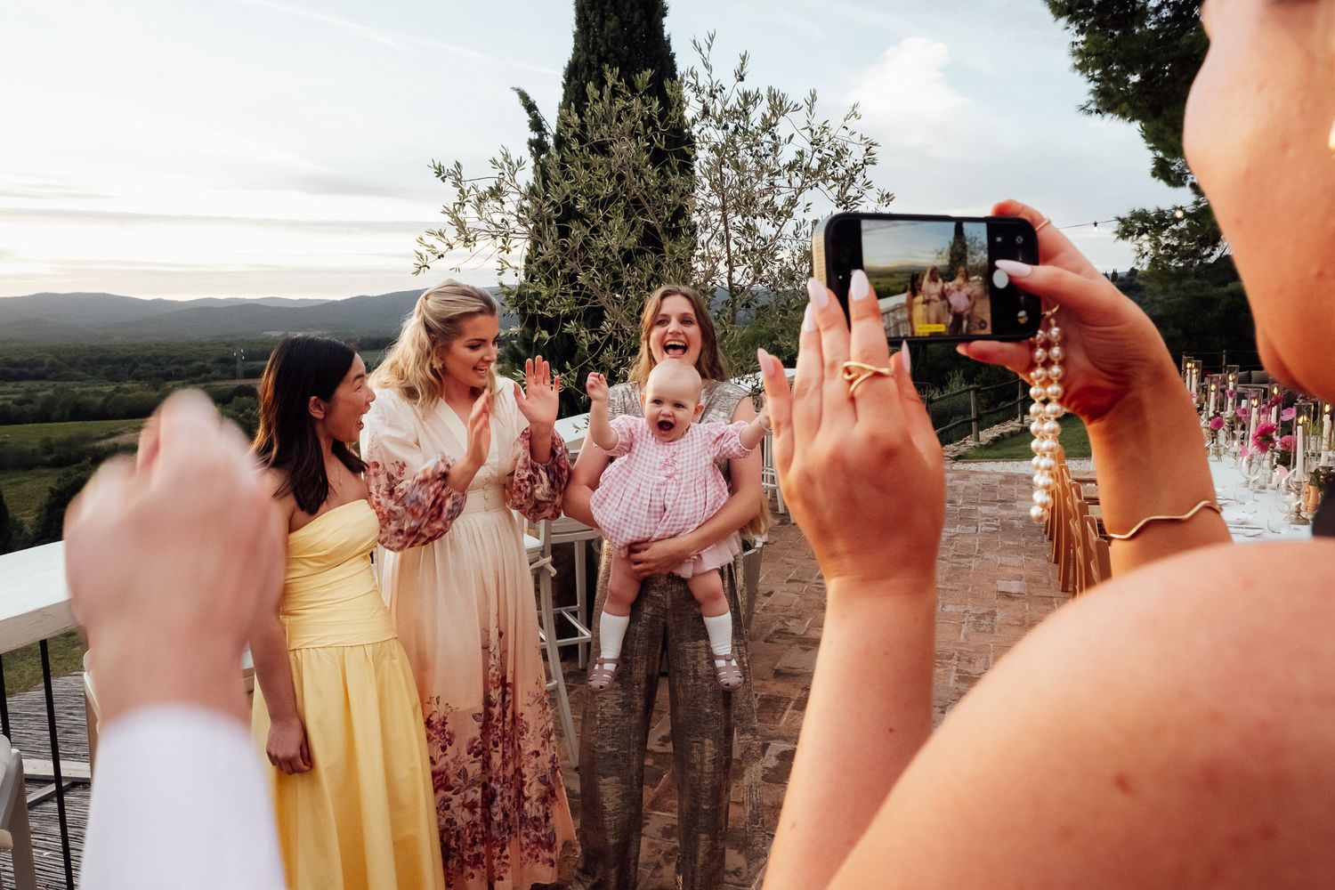 Guests in elegant dresses smile and clap around a baby at an outdoor celebration while another guest photographs the moment on a smartphone.