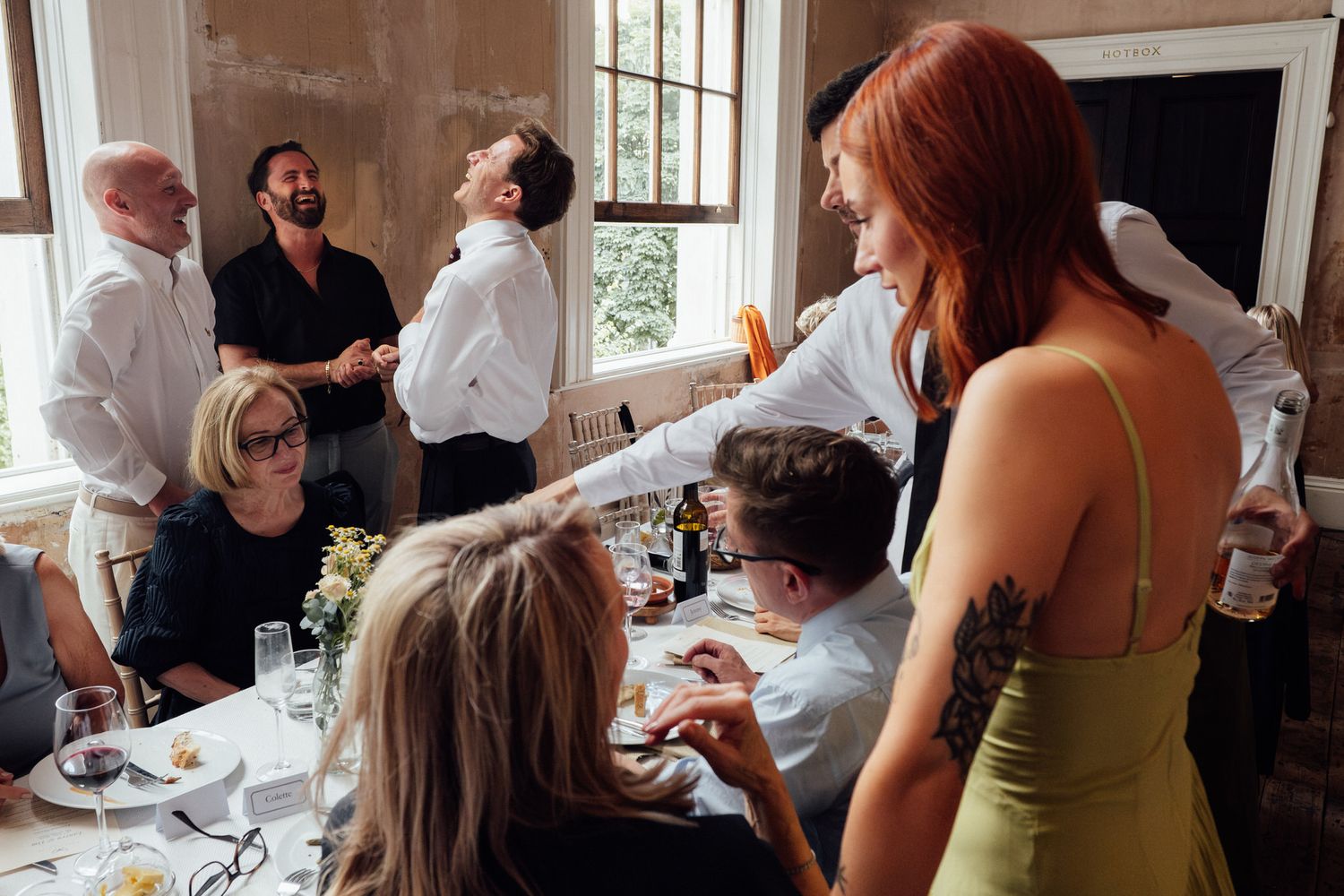 Wedding guests socializing at a reception dinner table, with a tattooed woman in a green dress gesturing.
