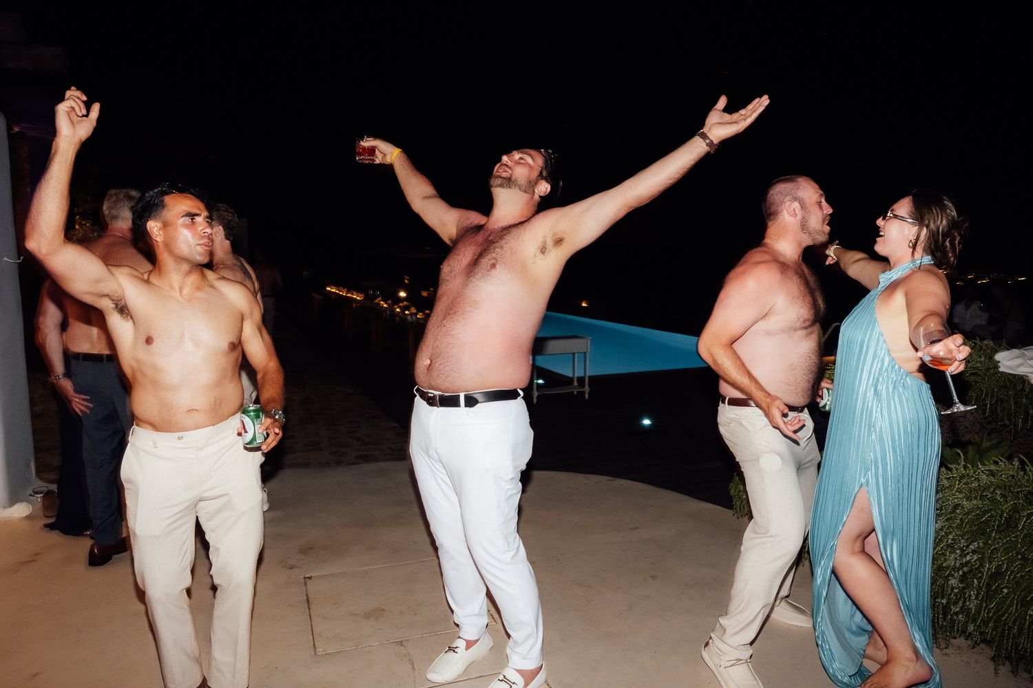 Shirtless men dancing outdoors at a night party near a pool, with a woman in a teal dress holding a drink.