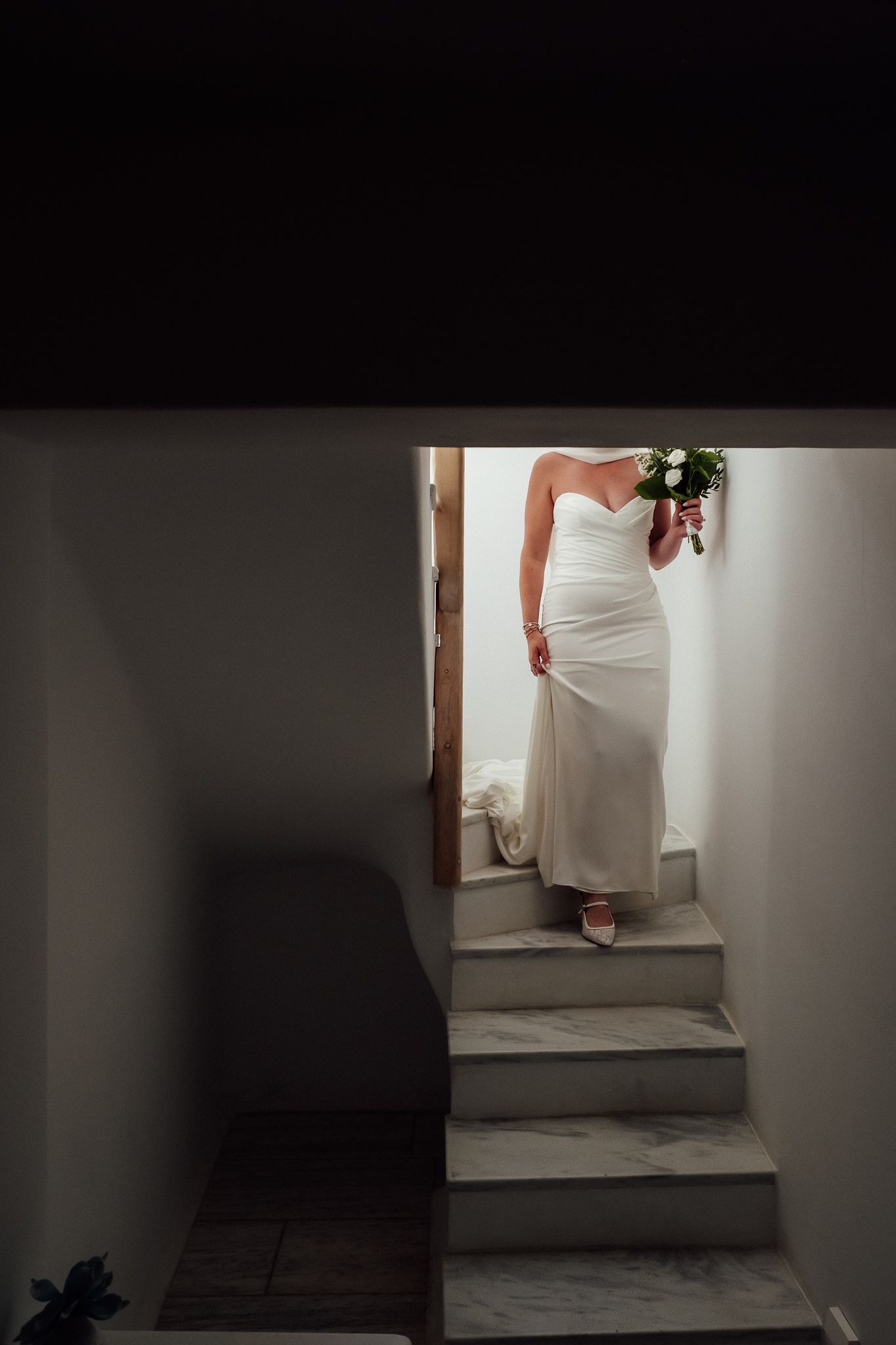 Bride in white strapless wedding gown holding bouquet, standing on marble stairs viewed through doorway.