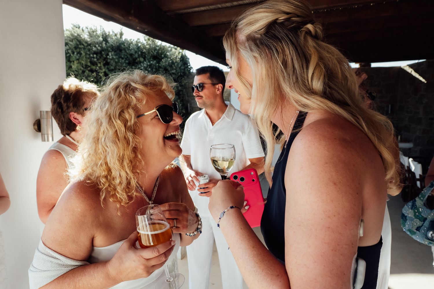 Two women laughing and toasting with wine glasses at an outdoor social gathering on a sunny day.