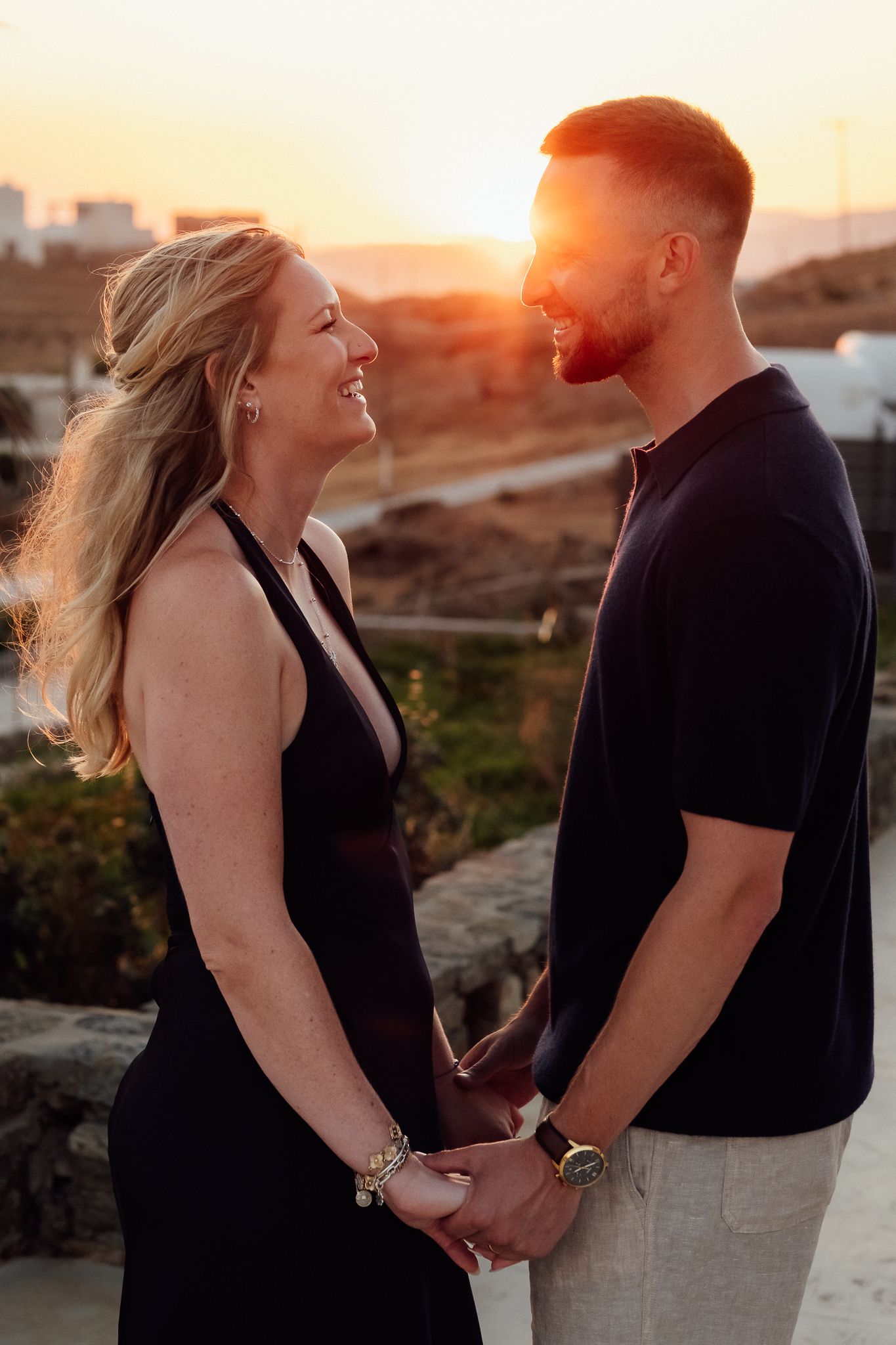 A couple holds hands and smiles at sunset, woman in black halter dress, man in navy tee, golden light behind them.