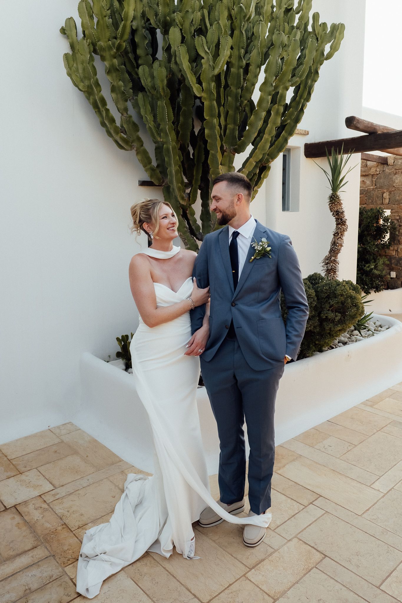Bride in white gown and groom in blue suit pose together in front of a large cactus at an outdoor wedding venue.