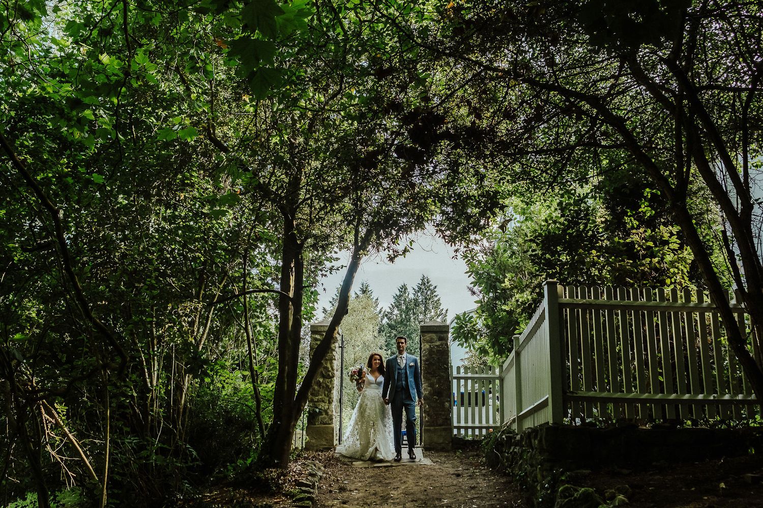 Wedding couple walks through a wooded path under natural green canopy.