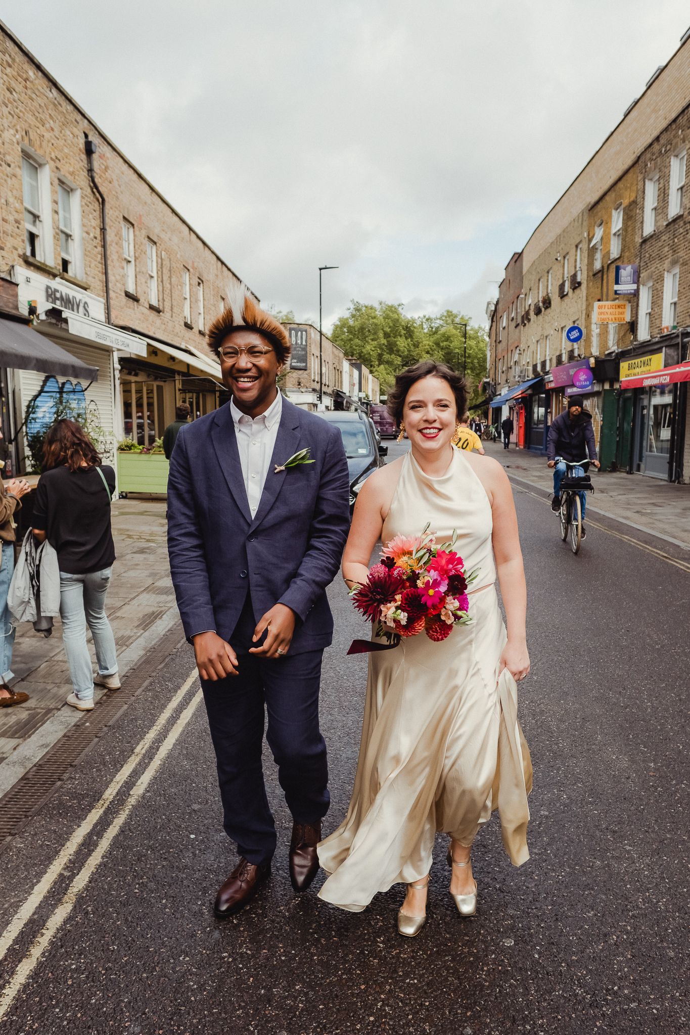A couple walking down an urban street with brick buildings on a cloudy day, one wearing a wedding dress with a bouquet.