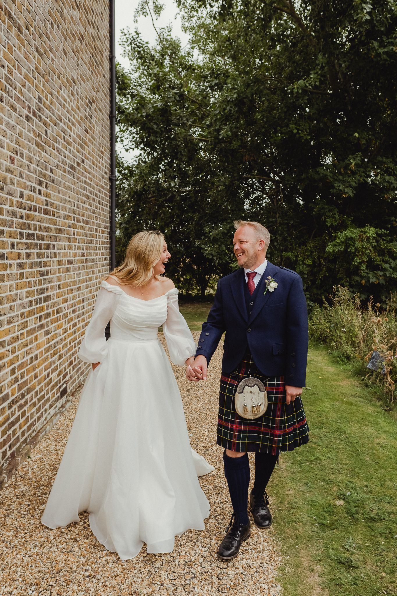 A couple in wedding attire walks along a brick wall and grassy path, with the groom wearing a traditional Scottish kilt.
