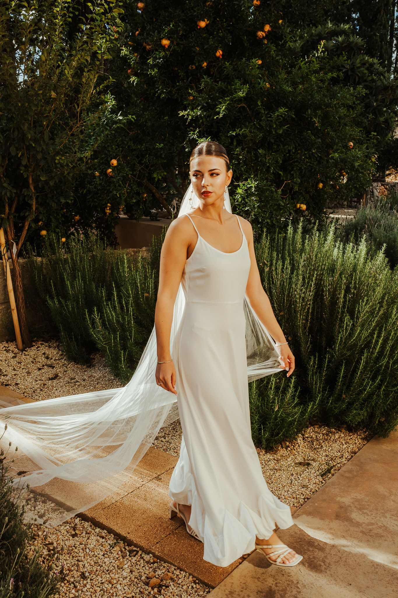 A simple white wedding dress with spaghetti straps and flowing veil flows in the breeze against a garden backdrop.