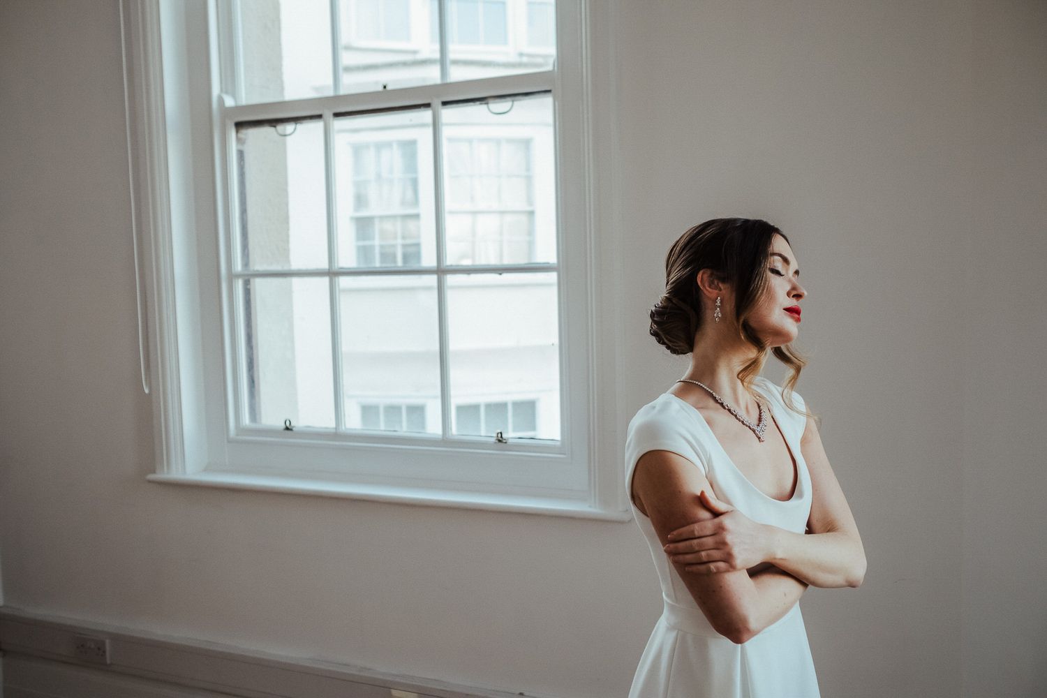 A person in a white dress stands thoughtfully by a bright window in a minimalist room.