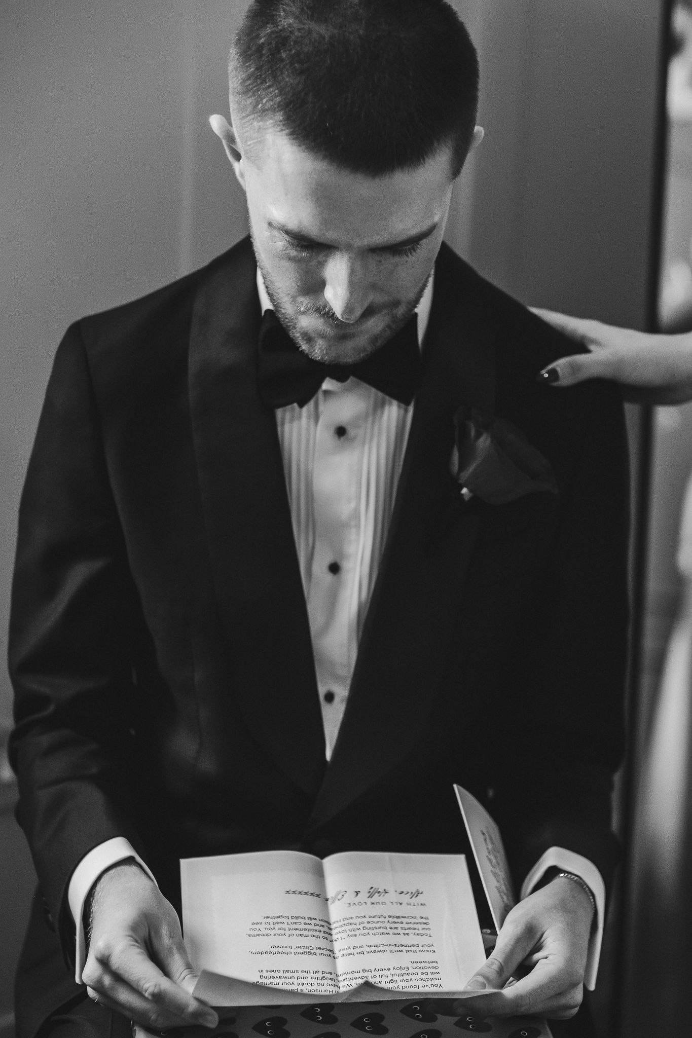 A groom in a black tuxedo and bow tie reads from a book during a wedding ceremony, captured in black and white.