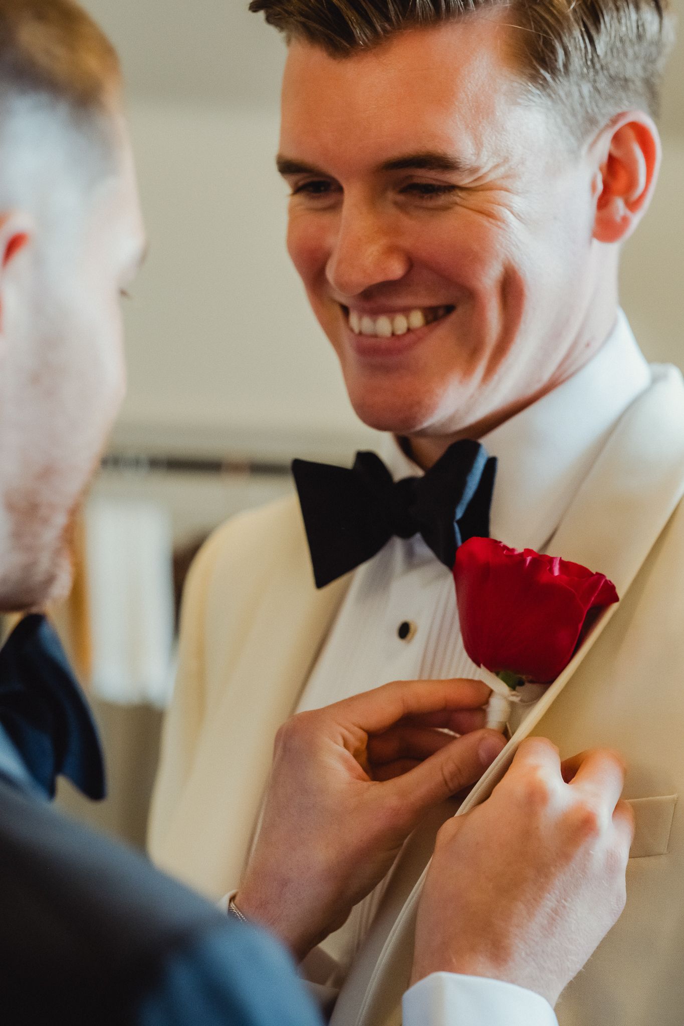 A person in a white tuxedo jacket and black bowtie has a red rose boutonniere pinned to their lapel by someone else.