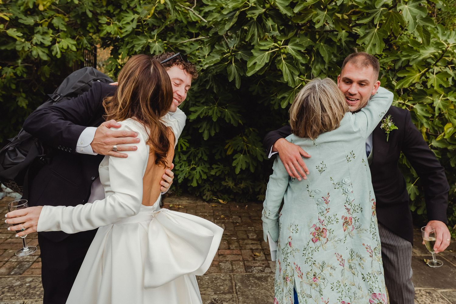 Two couples share joyful embraces outdoors against a backdrop of green foliage.