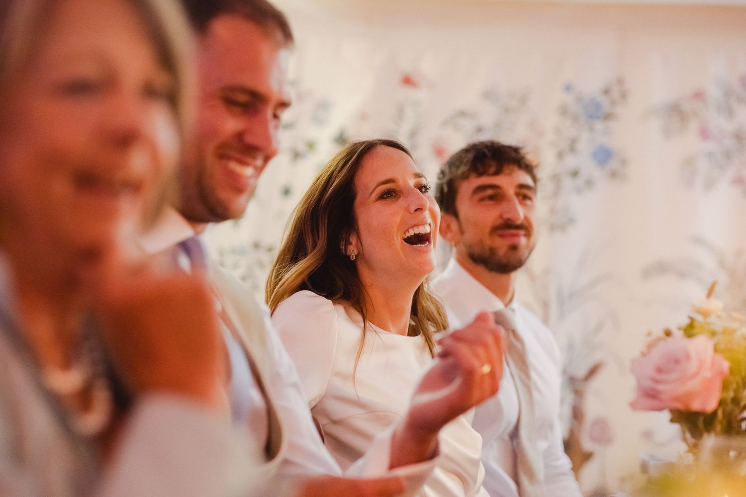 Joyful wedding guests laughing and celebrating at an outdoor reception with pink and blue floral decorations.