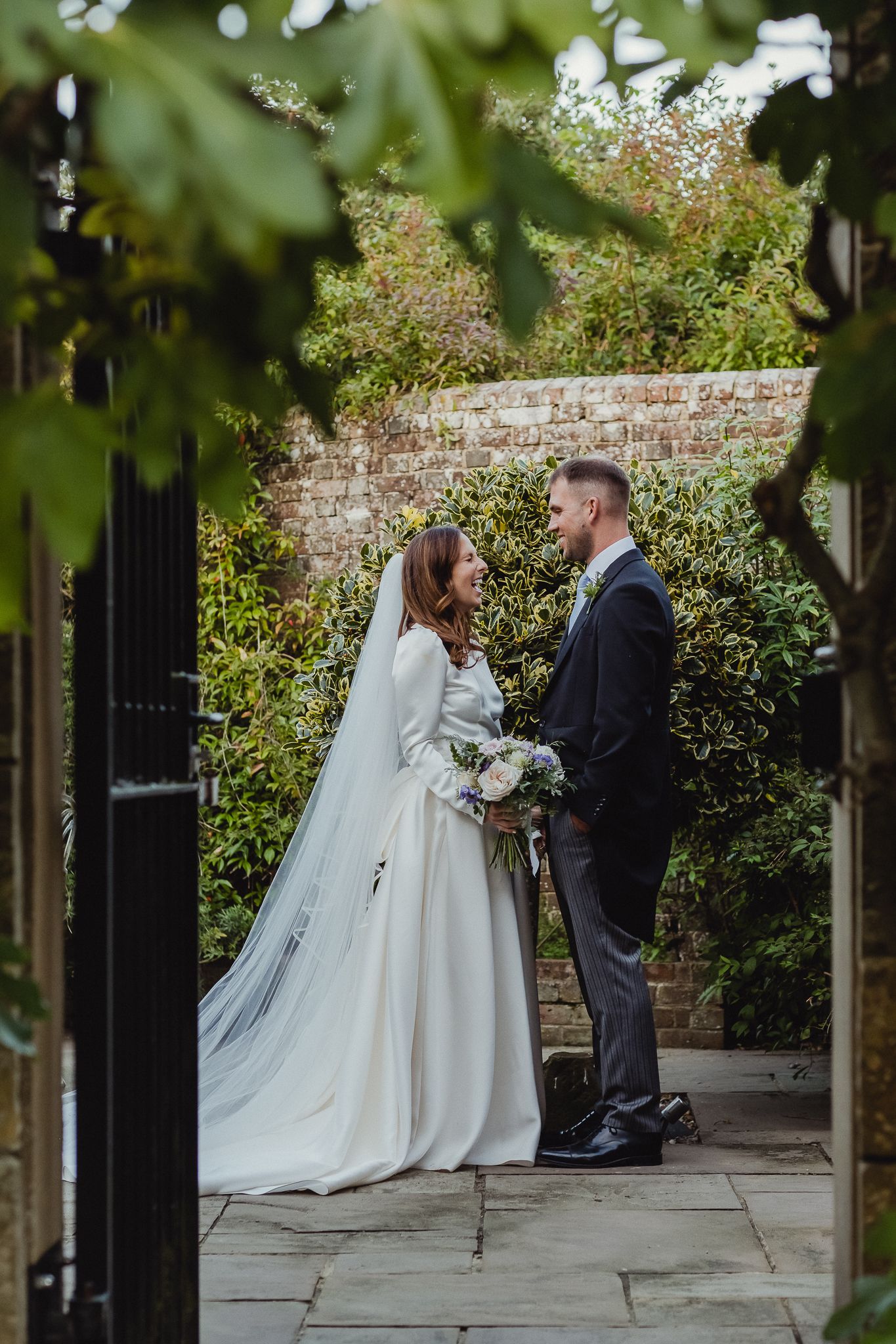 A couple shares an intimate moment in their wedding attire by a brick garden wall with lush greenery.