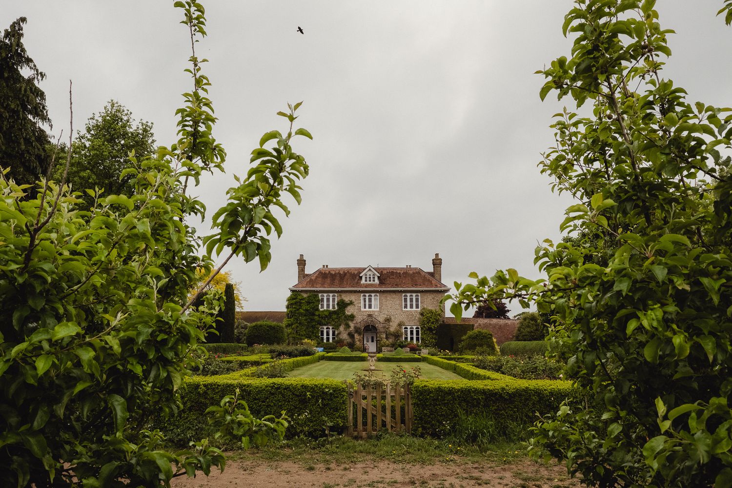 A red brick farmhouse sits amid lush green trees on an overcast day in the countryside.