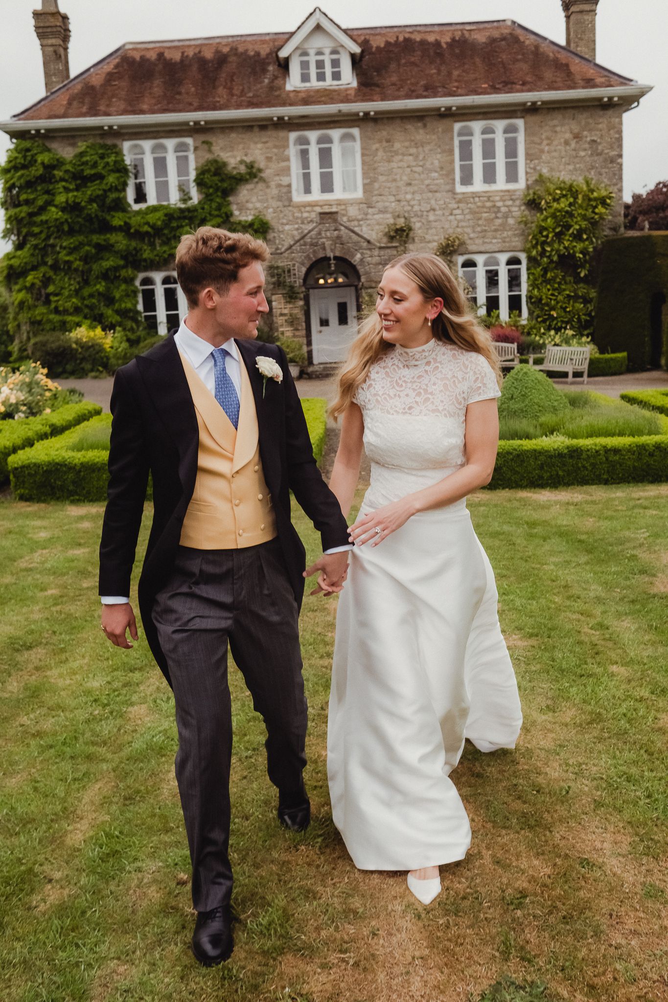 A couple in wedding attire walking and laughing together on the lawn of a stone country estate on a sunny day.