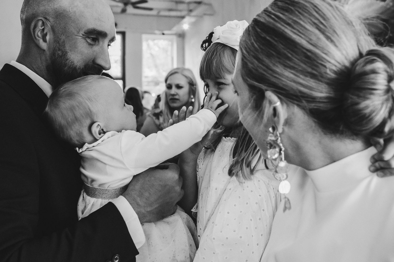 Black and white emotional moment captured at celebration as baby reaches out to touch in candid expression of joy.