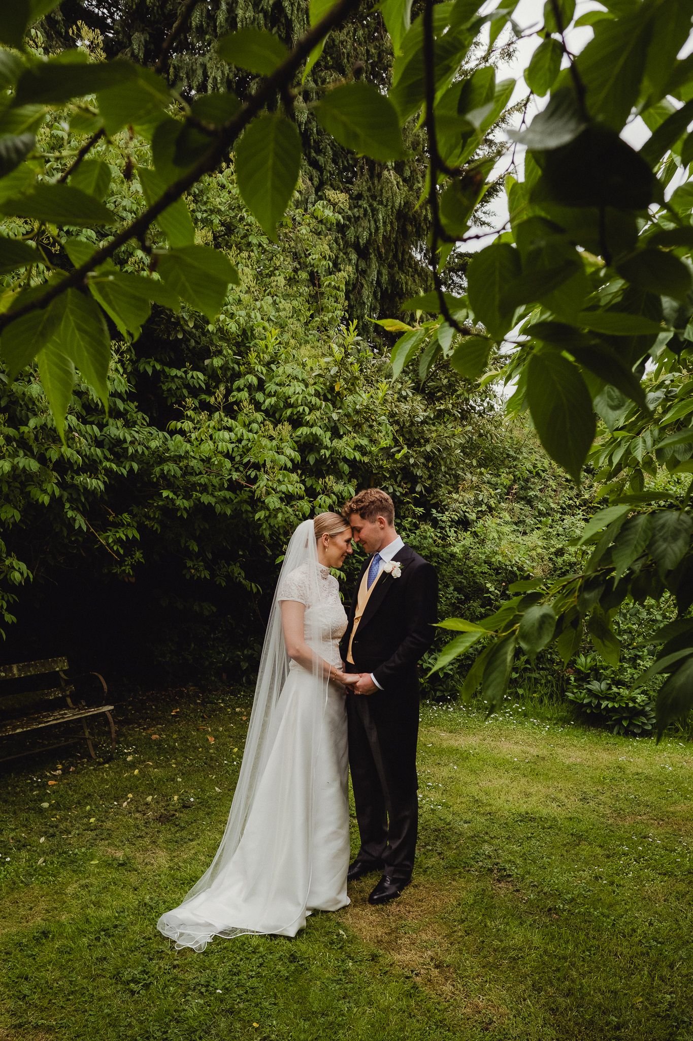 A couple in formal wedding attire embrace in a lush green garden setting during their outdoor ceremony.