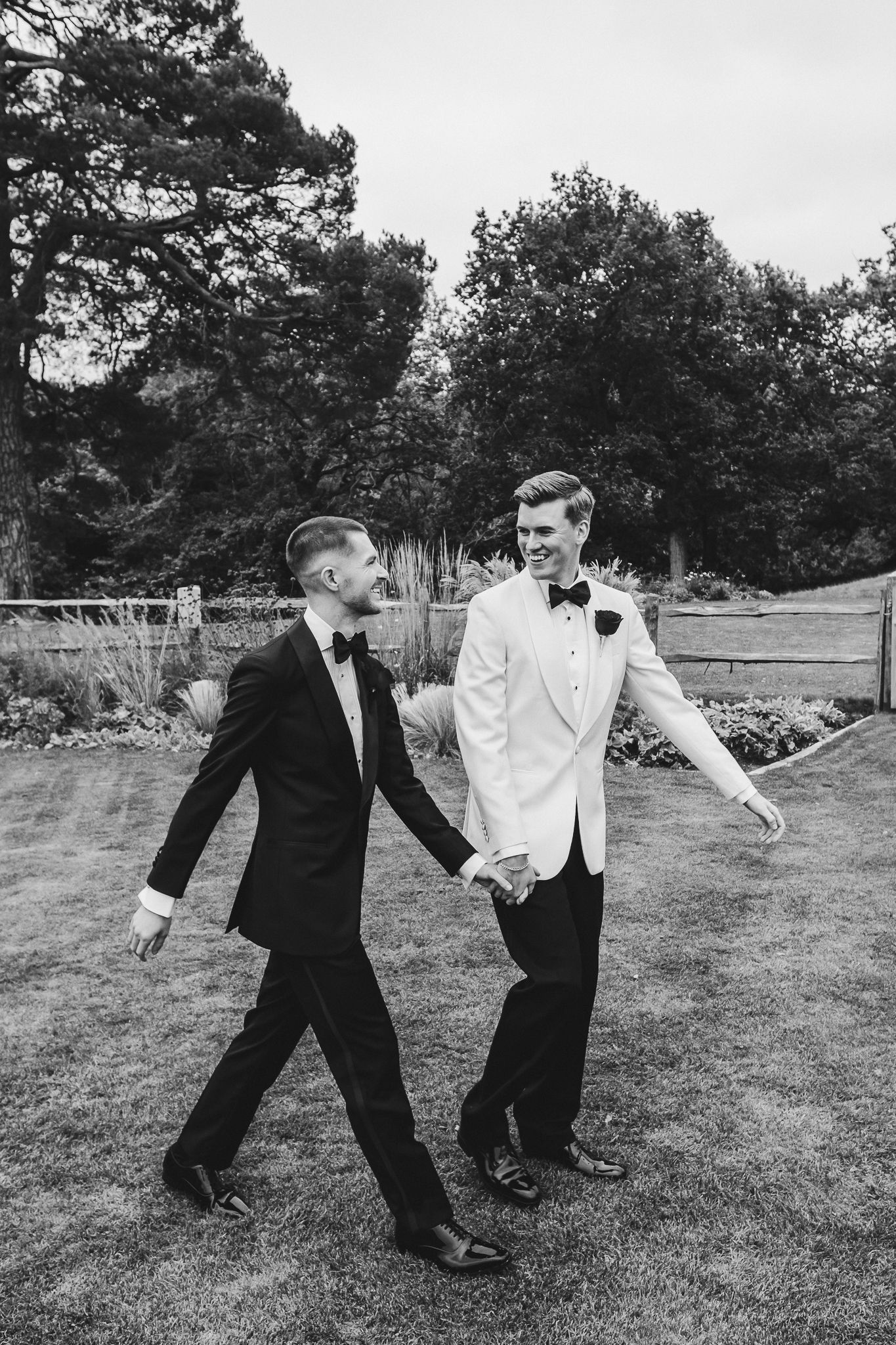 Two men in formal attire playfully running and dancing together on a grassy lawn in black and white photography.