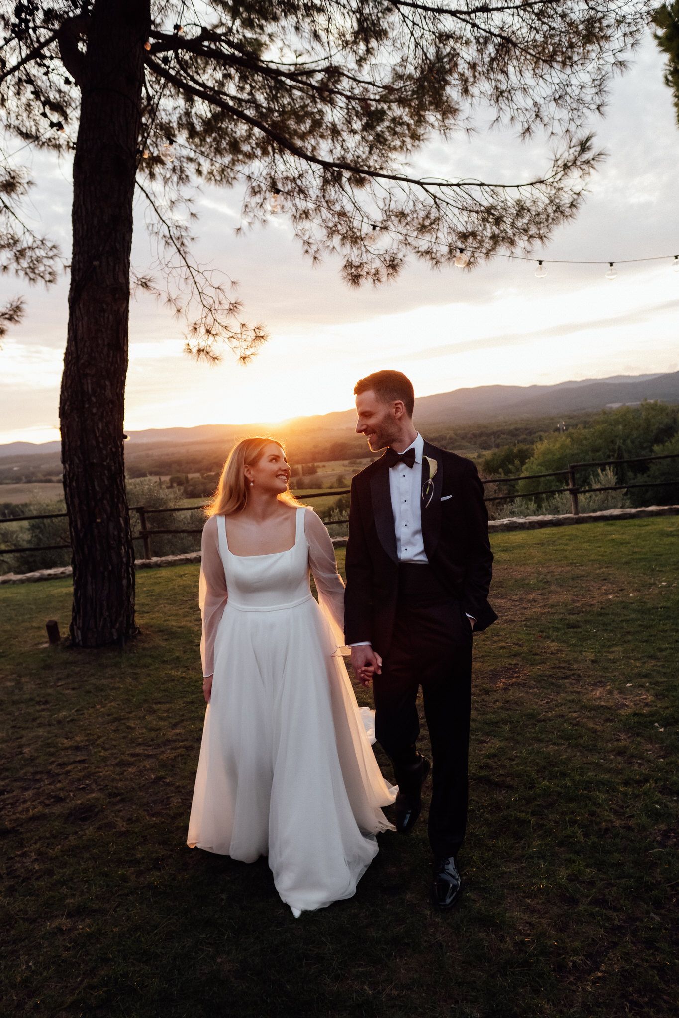 Bride in a white gown and groom in a black tuxedo hold hands and smile at each other under a tree at sunset in the countryside.