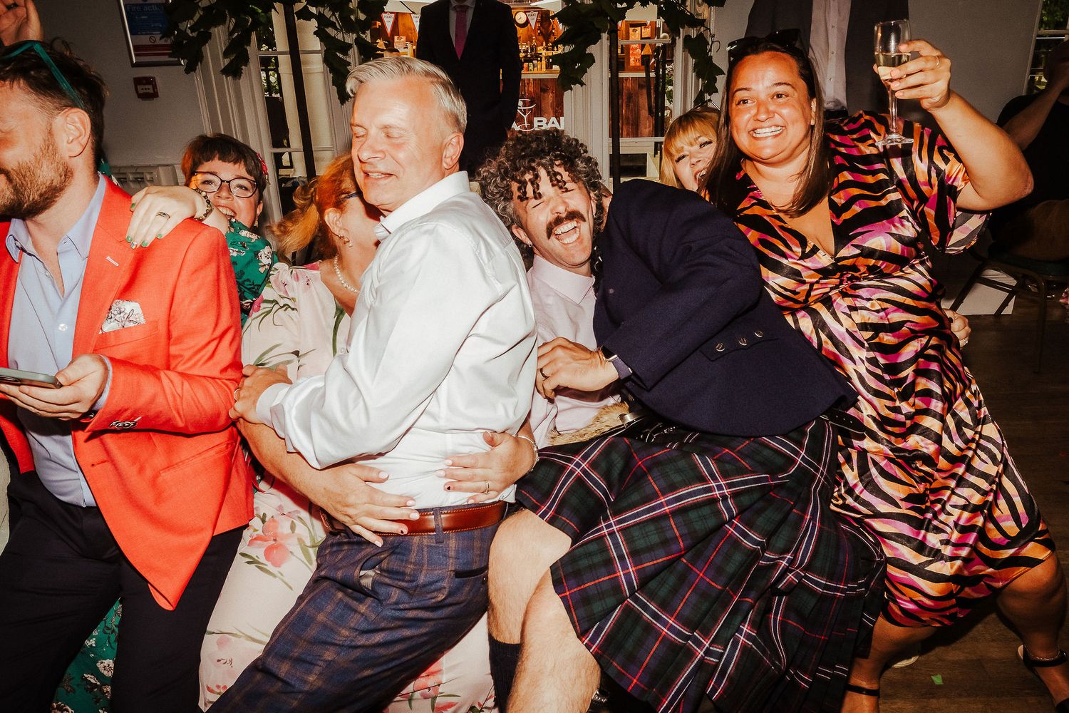 Guests in colorful outfits, including plaid kilts and an orange blazer, laugh and dance together at a lively wedding celebration.