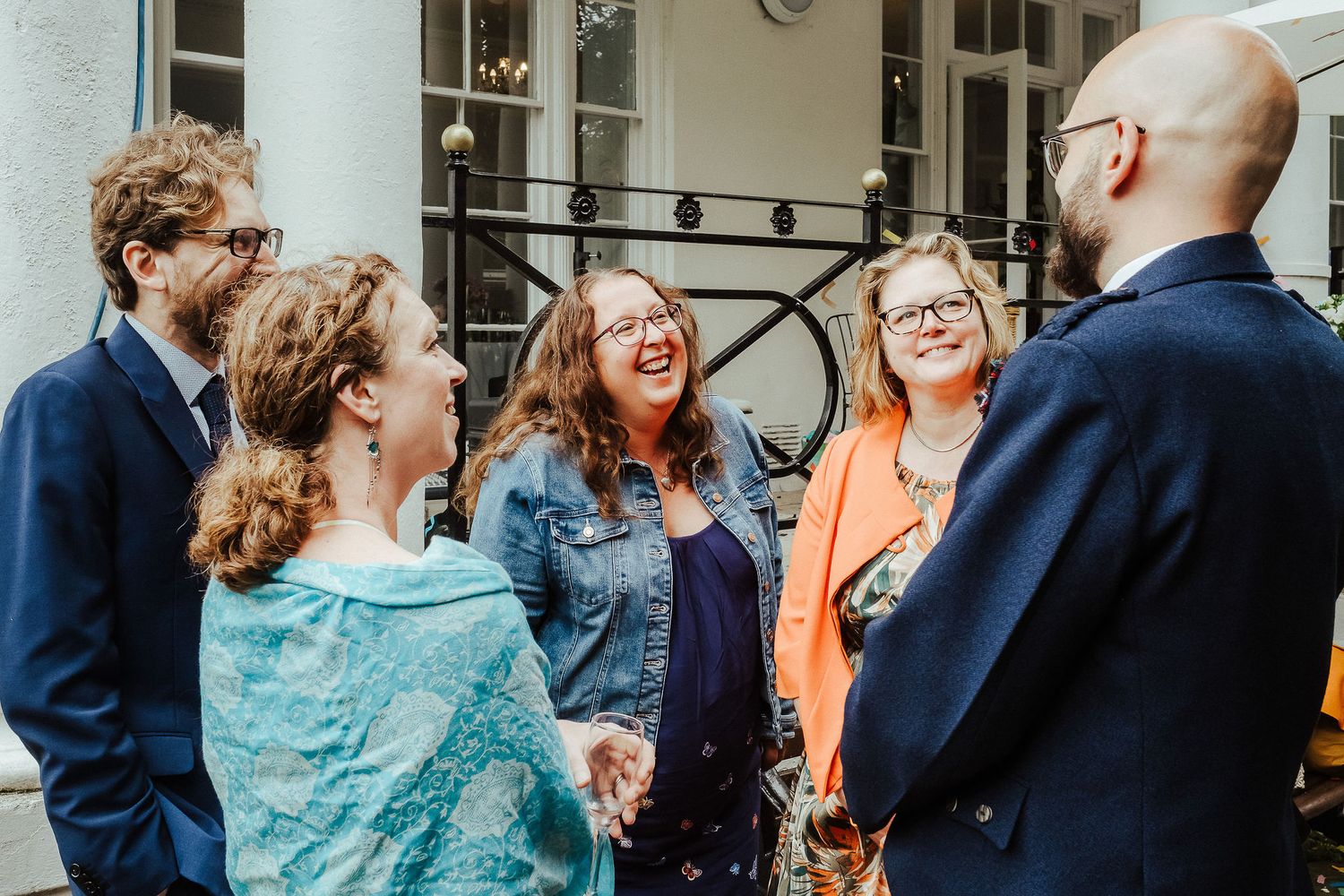 Five smiling friends in dressy outfits chat and laugh together outside a historic building with wrought iron railings.