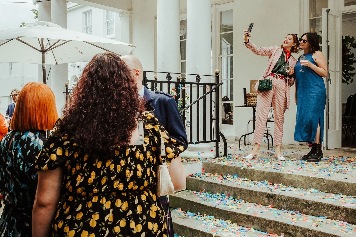 Wedding guests in colorful dresses stand on stone steps covered in confetti while two women smile and take photos outside a white building.