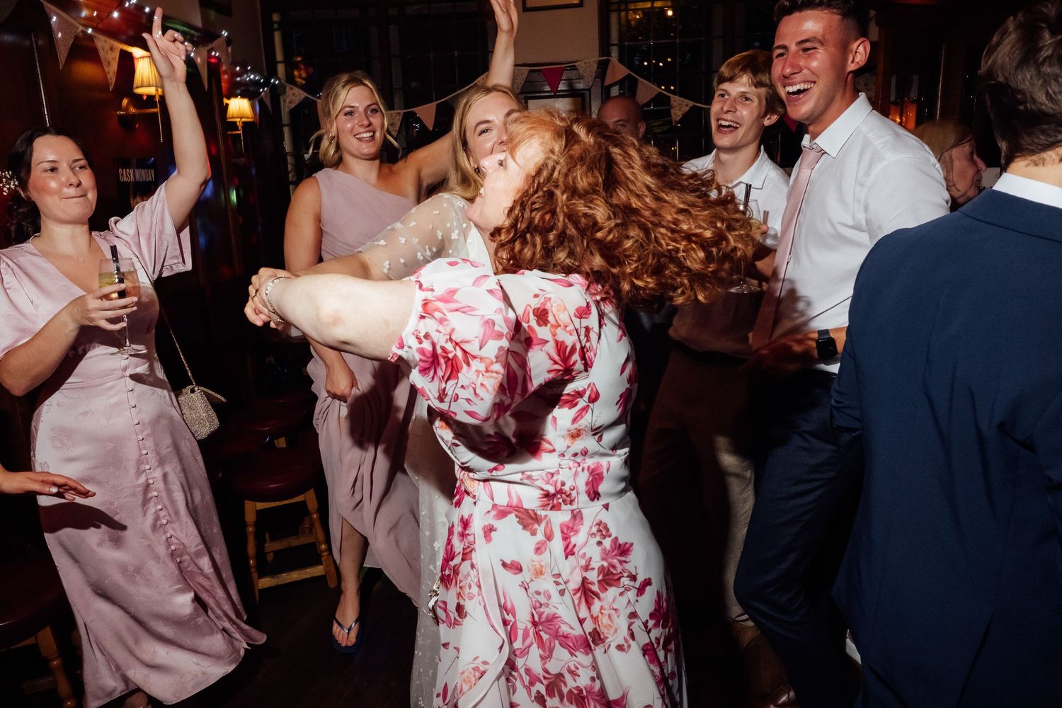 Guests in floral and pastel dresses dance joyfully with raised arms at a lively indoor party or wedding reception.