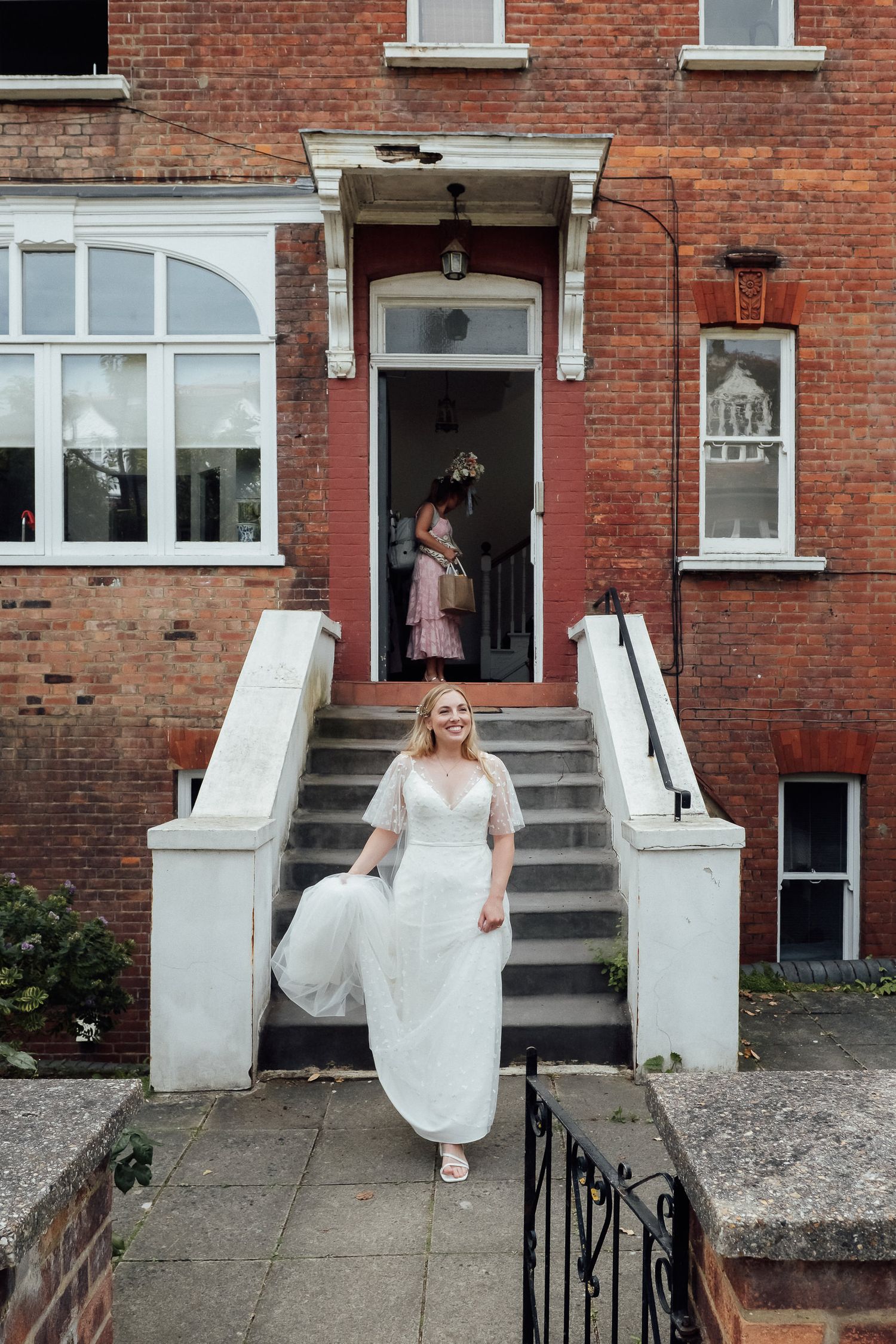 Bride in a white gown with sheer sleeves descends stone steps from a red brick house while a woman in a pink dress stands in the doorway.