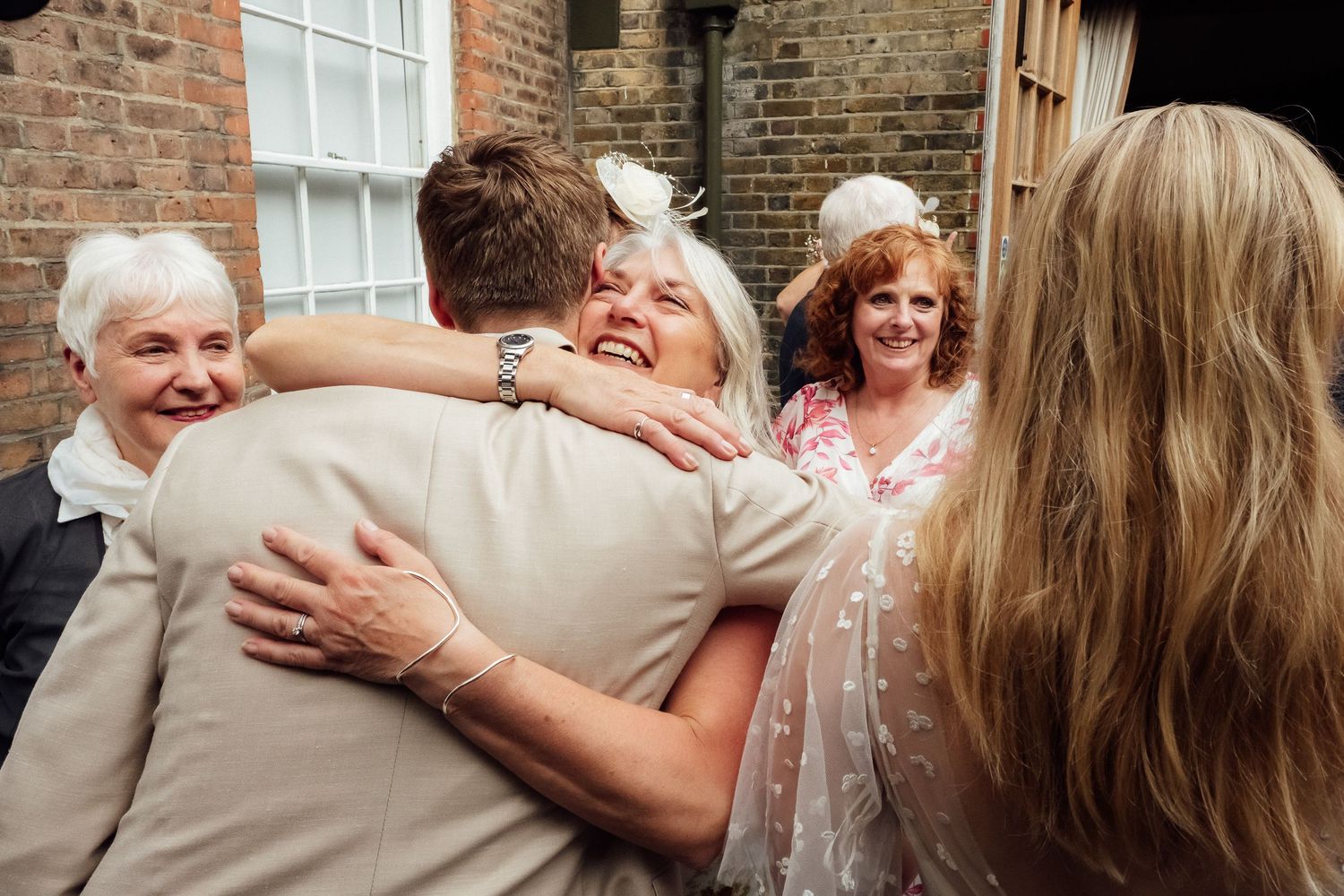 Older woman smiles warmly as she hugs a man in a beige jacket while other guests look on at a joyful gathering outside a brick building.