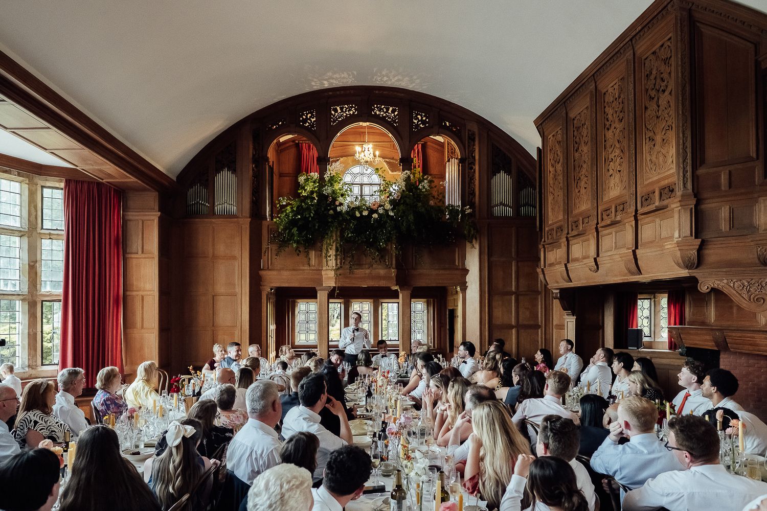 Elegant wedding reception in a grand oak-paneled hall with vaulted ceiling, floral chandelier, and guests seated at long tables.