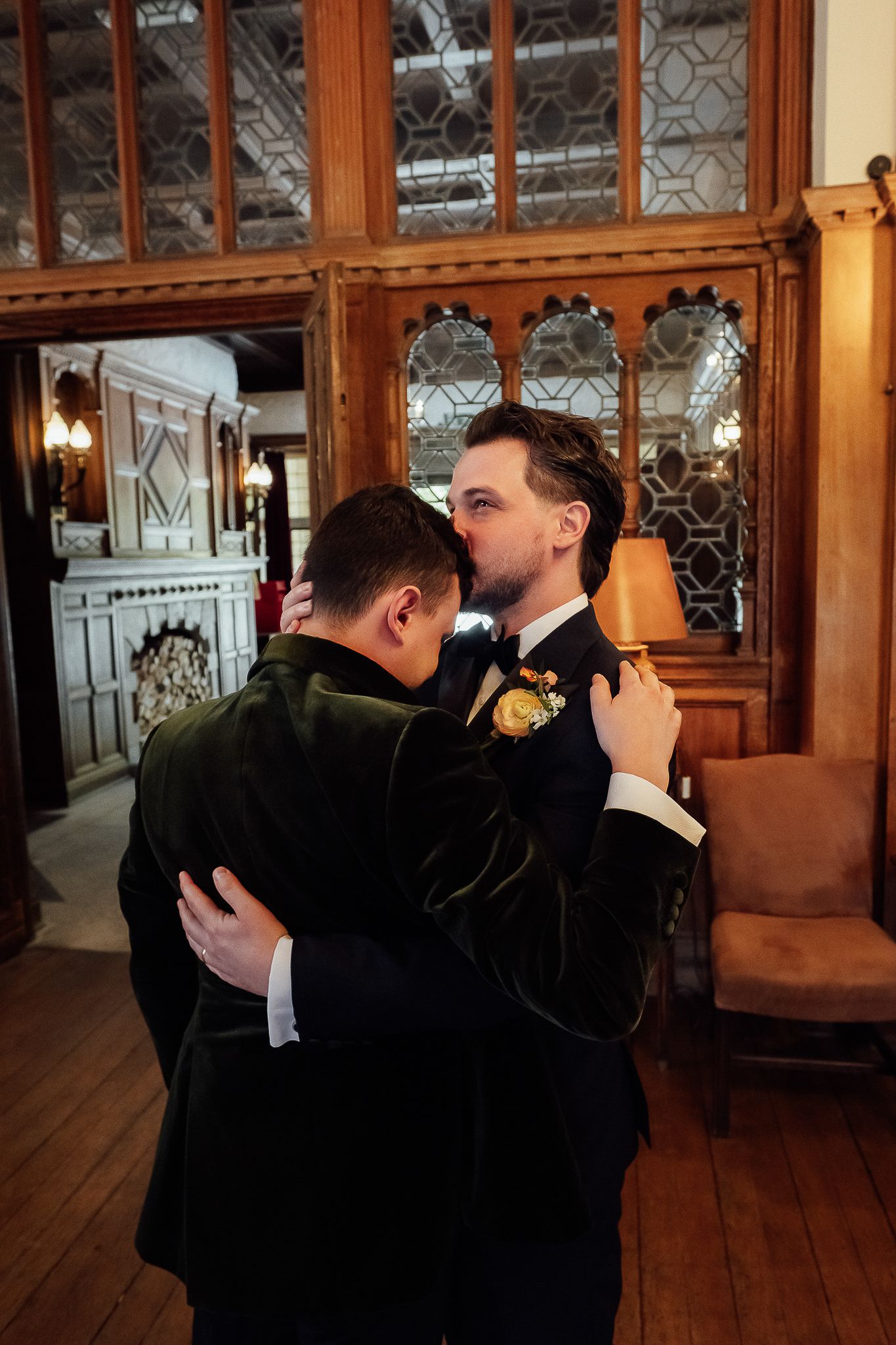 Two grooms share a tender embrace in an elegant wood-paneled room during their wedding celebration.