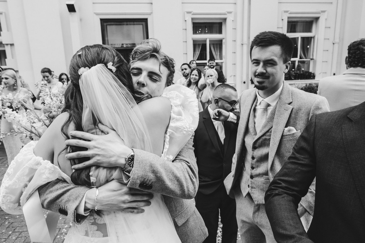 A joyful embrace between wedding guests captured in black and white as others look on during an outdoor celebration.