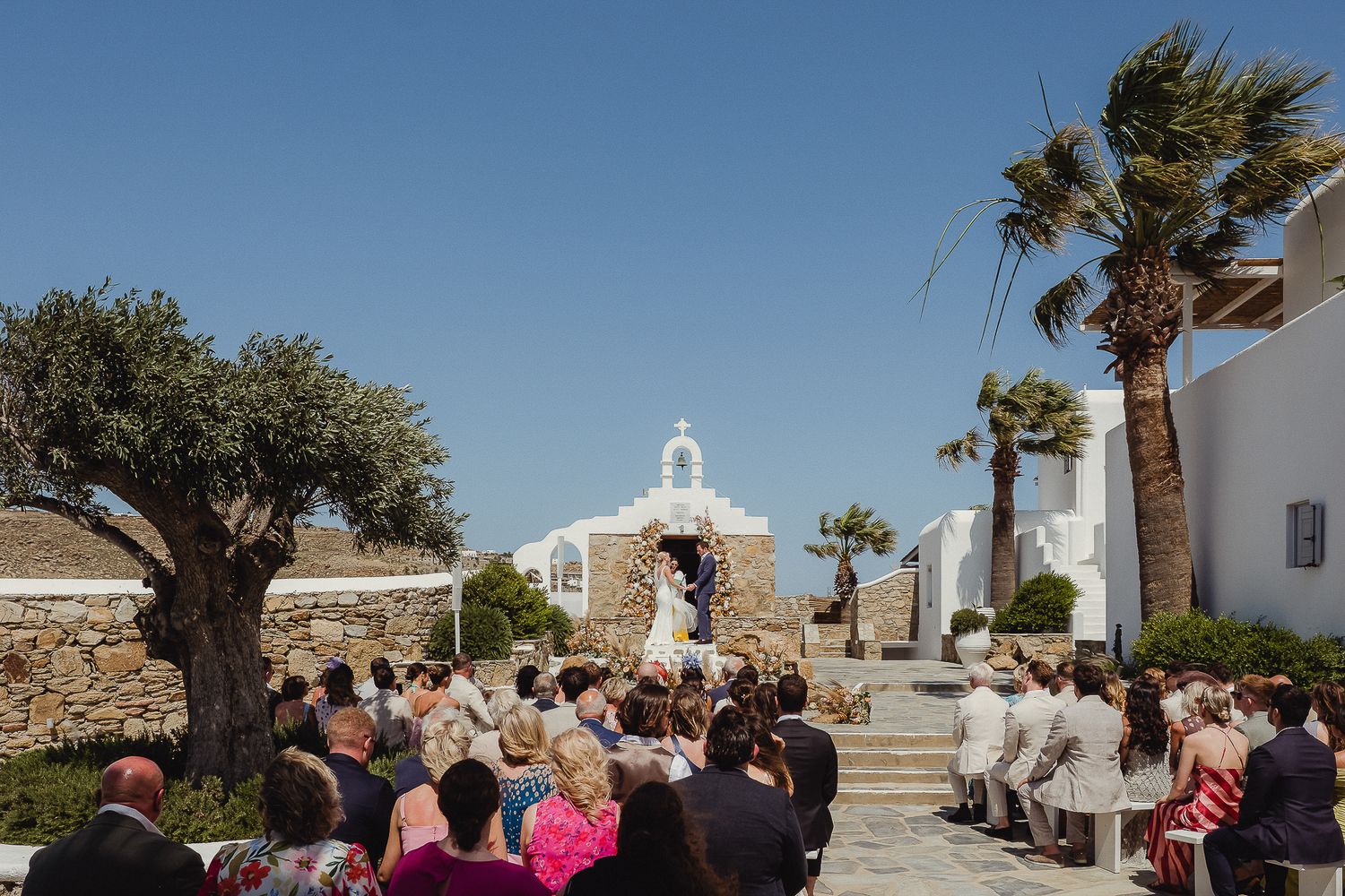 People gathered outside a white Mediterranean church with palm trees during an outdoor ceremony under blue skies.